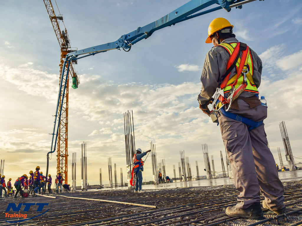 Construction workers on a building site with a crane and steel reinforcement bars, one worker in the foreground wearing a yellow safety helmet and harness, a sunset or sunrise sky in the background.