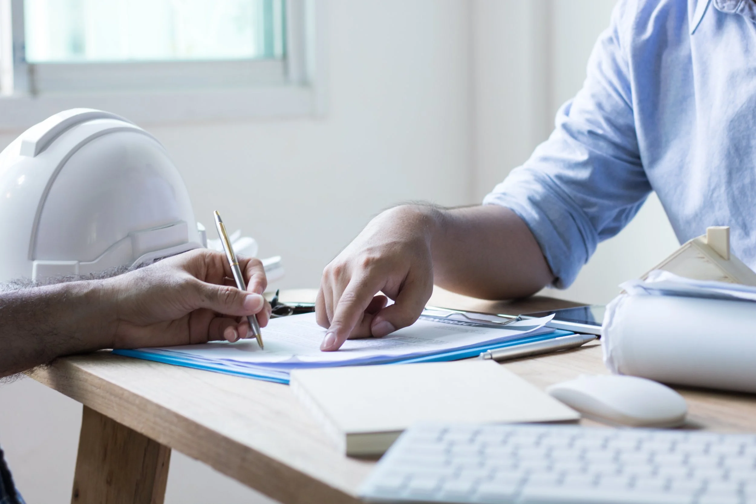 Two people discussing documents at a desk with a white helmet, rolled-up paper, and electronic devices.
