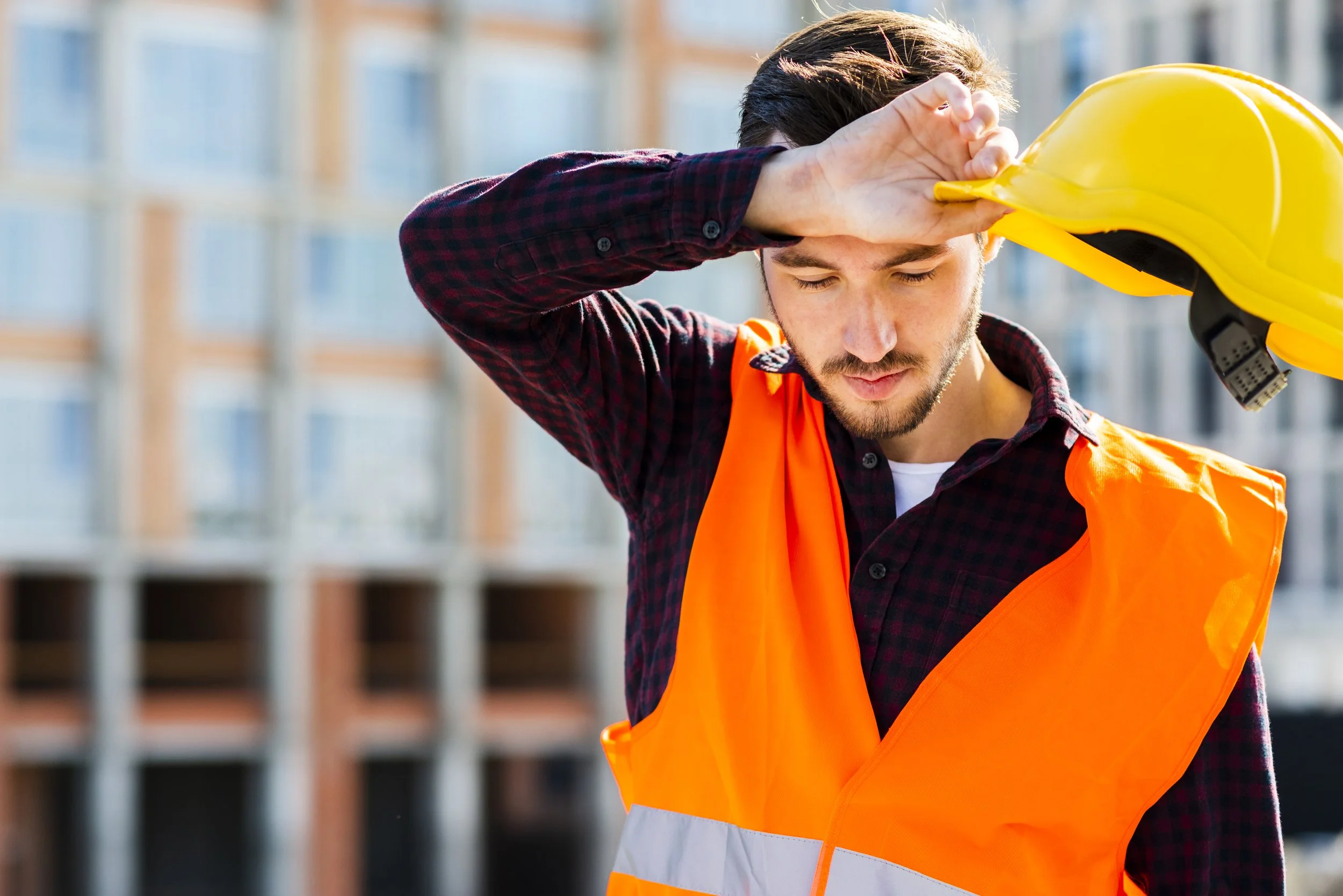 A construction worker with a beard and mustache wearing an orange safety vest, holding a yellow safety helmet, and saluting with his right hand on his forehead at a construction site.