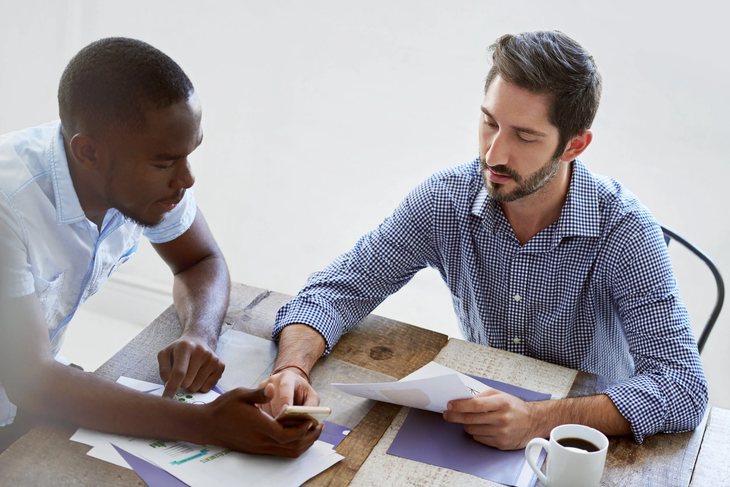 Two men working together at a wooden table, looking at documents and a smartphone, with a cup of coffee nearby.