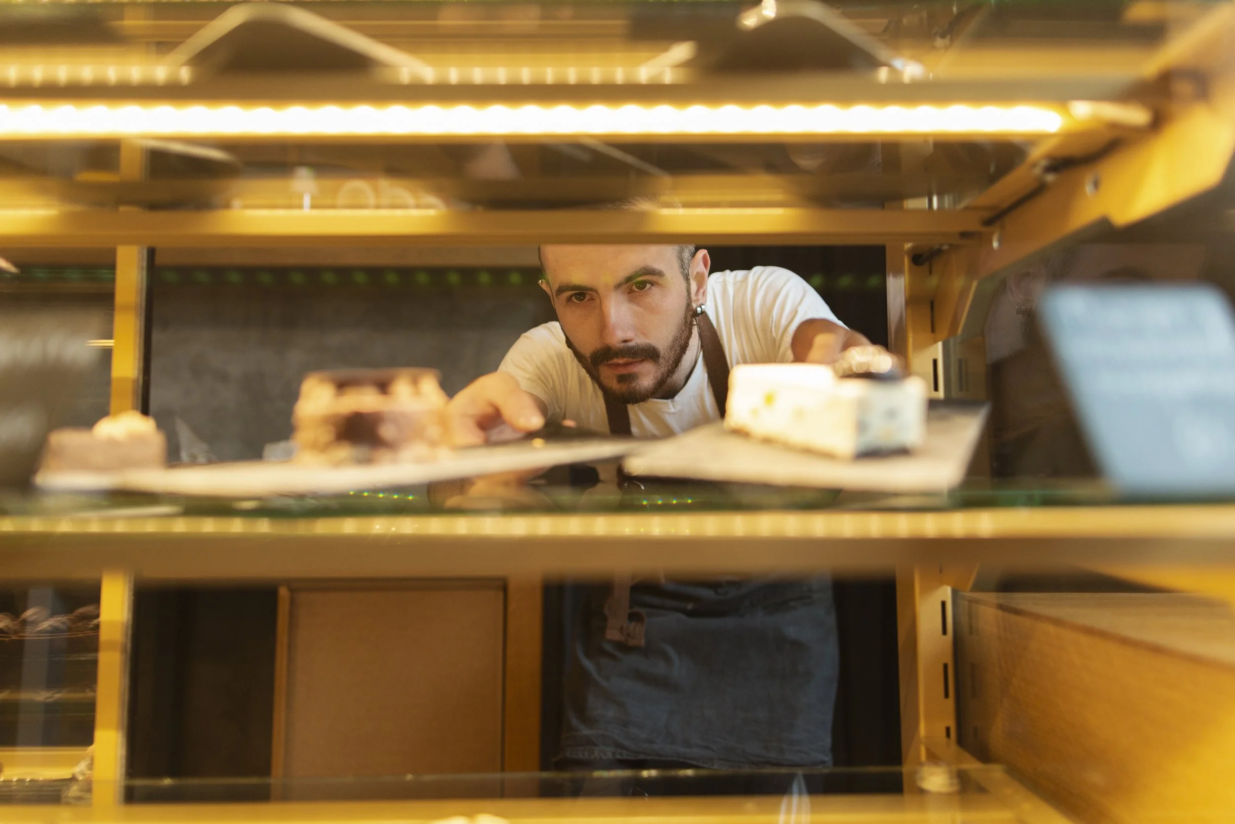 A man with a beard reaching into a bakery display case to pick out desserts. The photo is taken from inside the case, looking outward at the man.