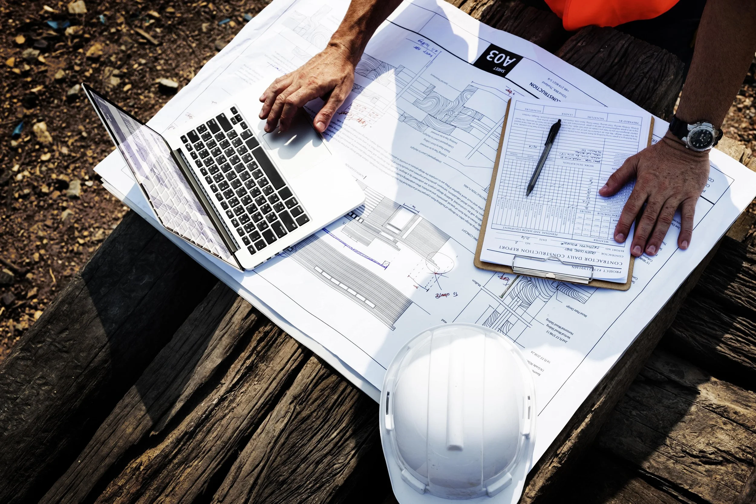 Architectural blueprints, a laptop, a clipboard with documents, a pen, and a white safety helmet on a wooden surface outdoors