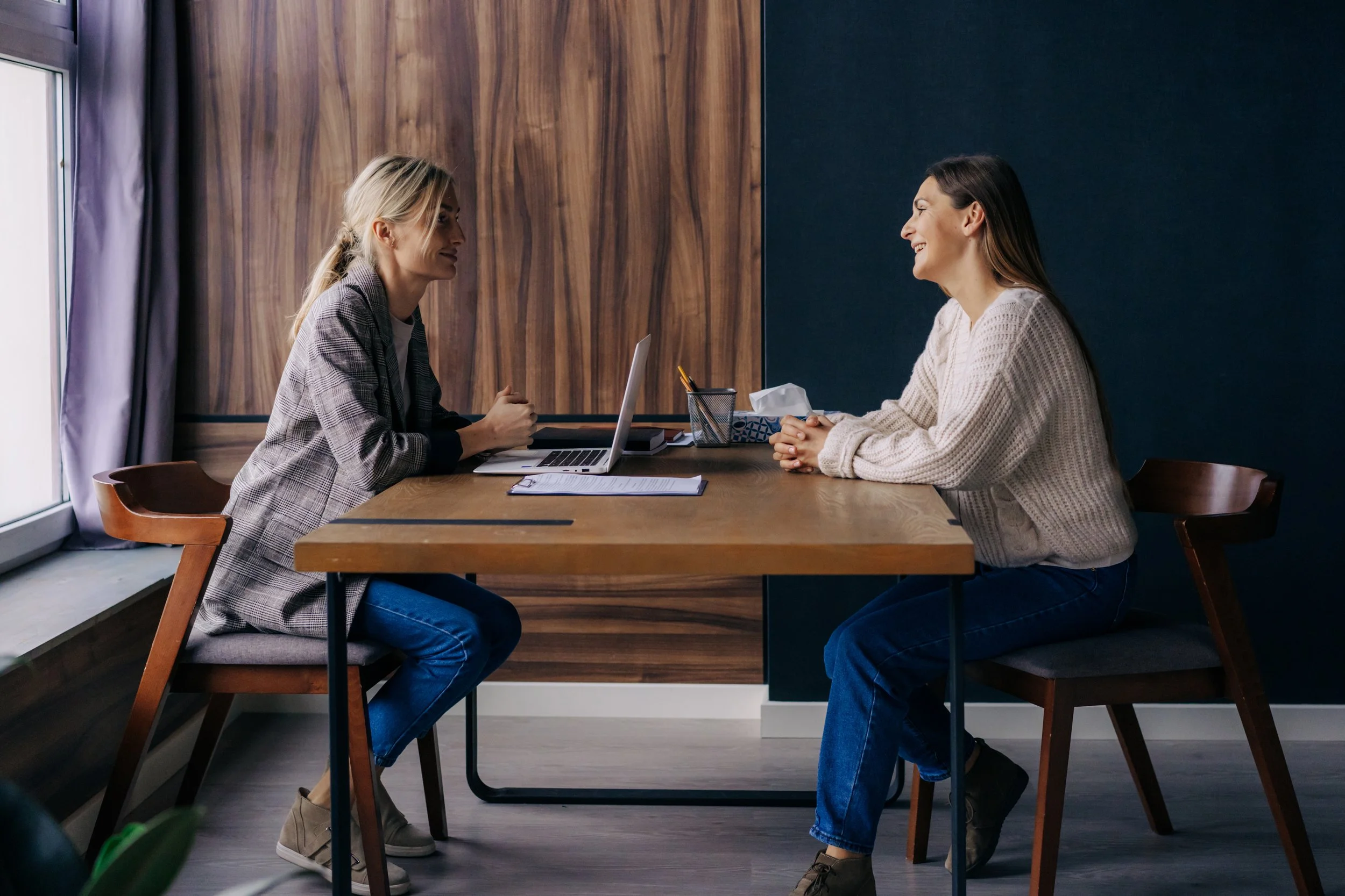 Two women having a conversation at a desk during an interview or meeting, in a room with wood-paneled walls and a large window.
