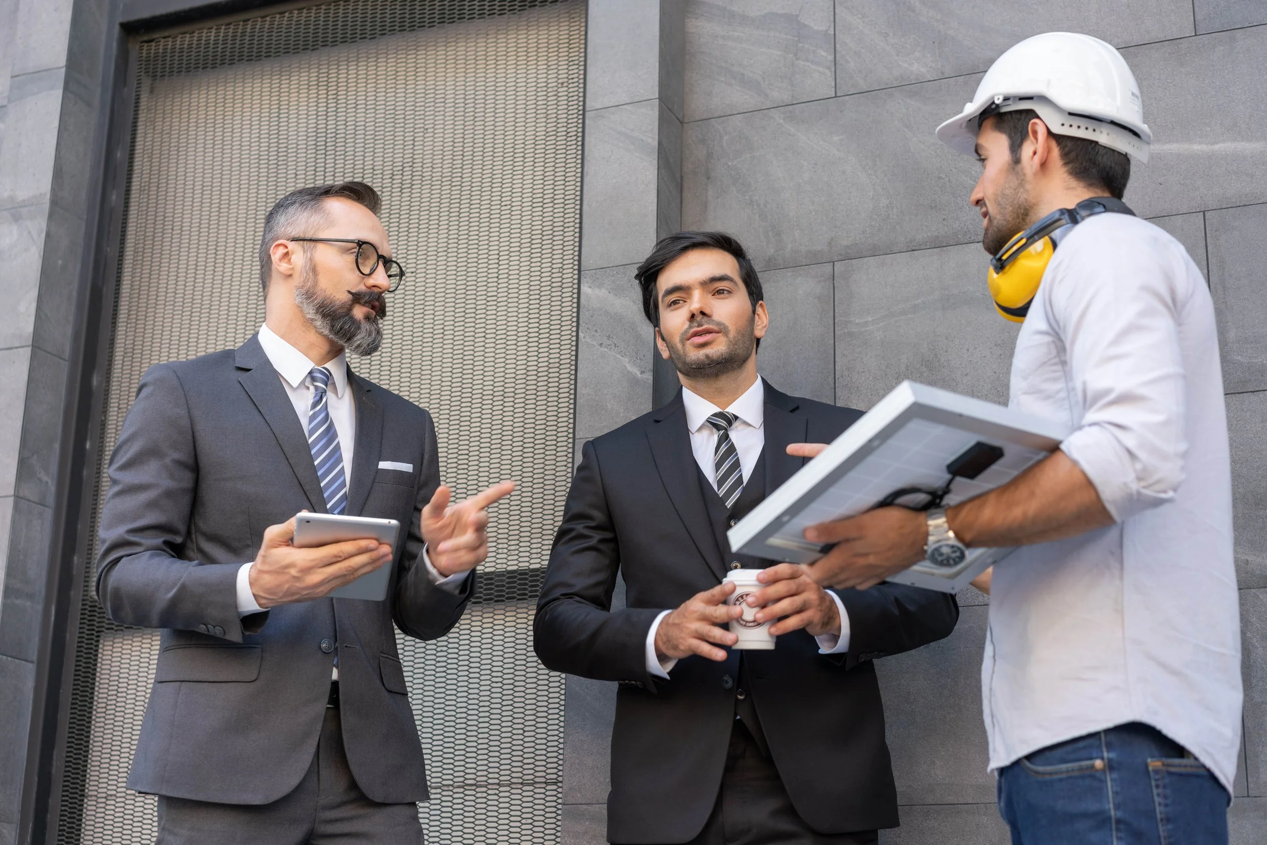 Three men, two in suits and one in casual work attire with a helmet and headphones, having a discussion outside a modern building.