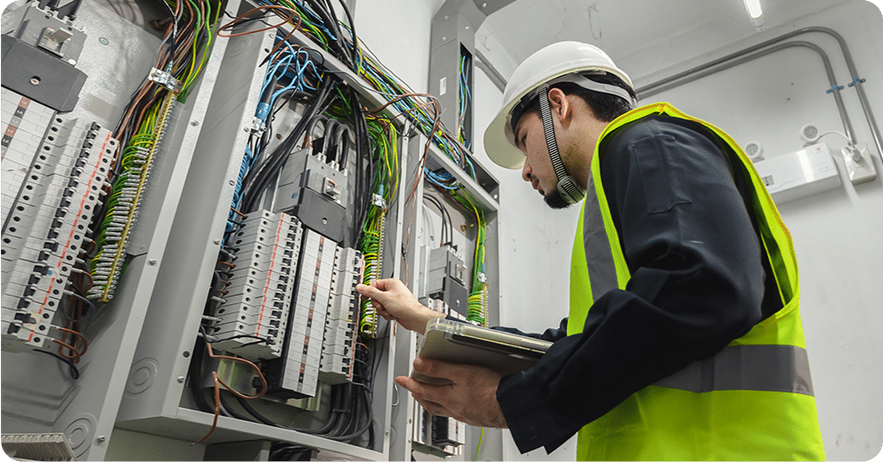 Electrician inspecting wiring and circuit breakers in an electrical panel, wearing a safety helmet and vest, holding a clipboard in a well-lit room.