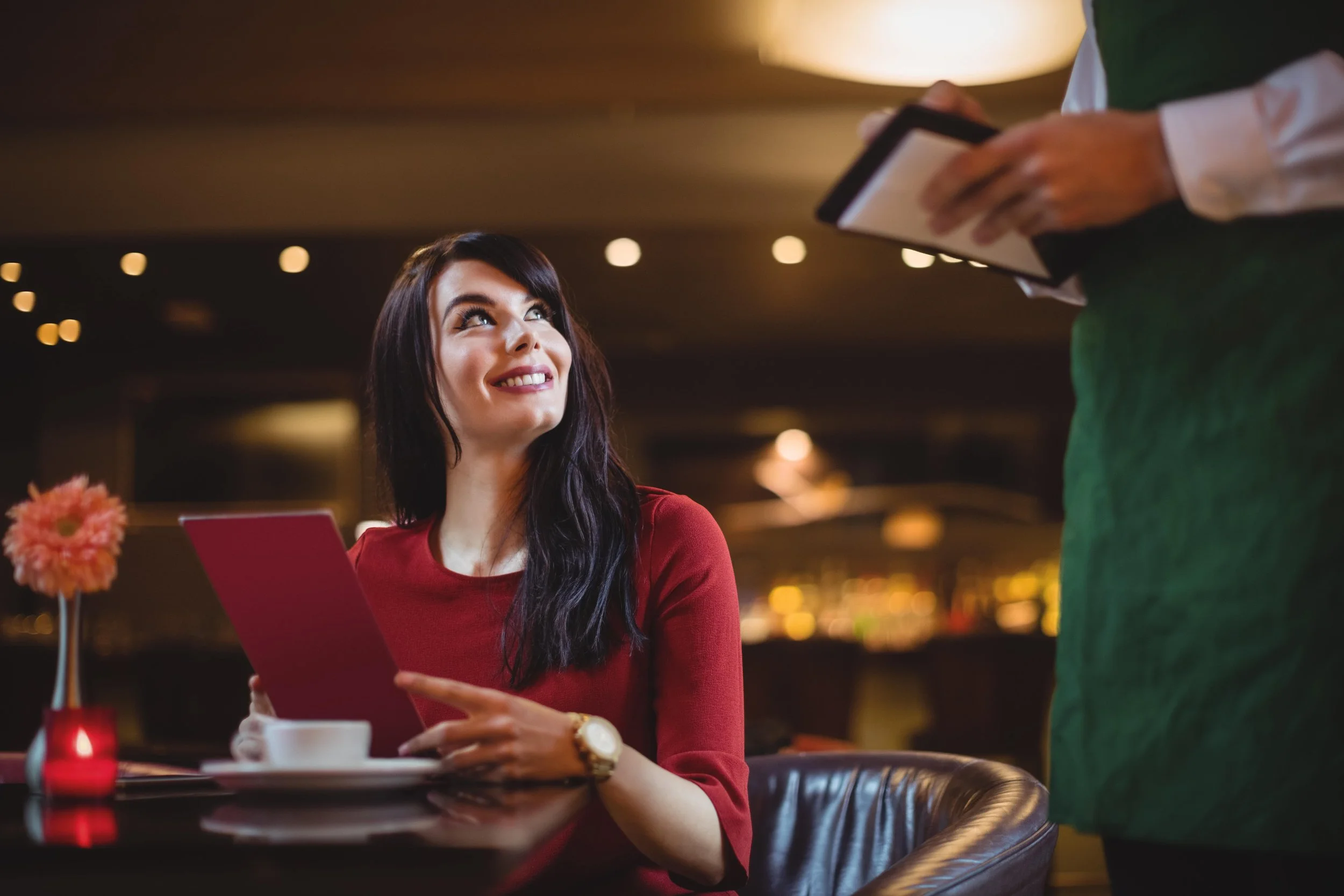 A woman in a red dress sitting at a restaurant table, holding a menu and smiling up at a server who is taking her order.