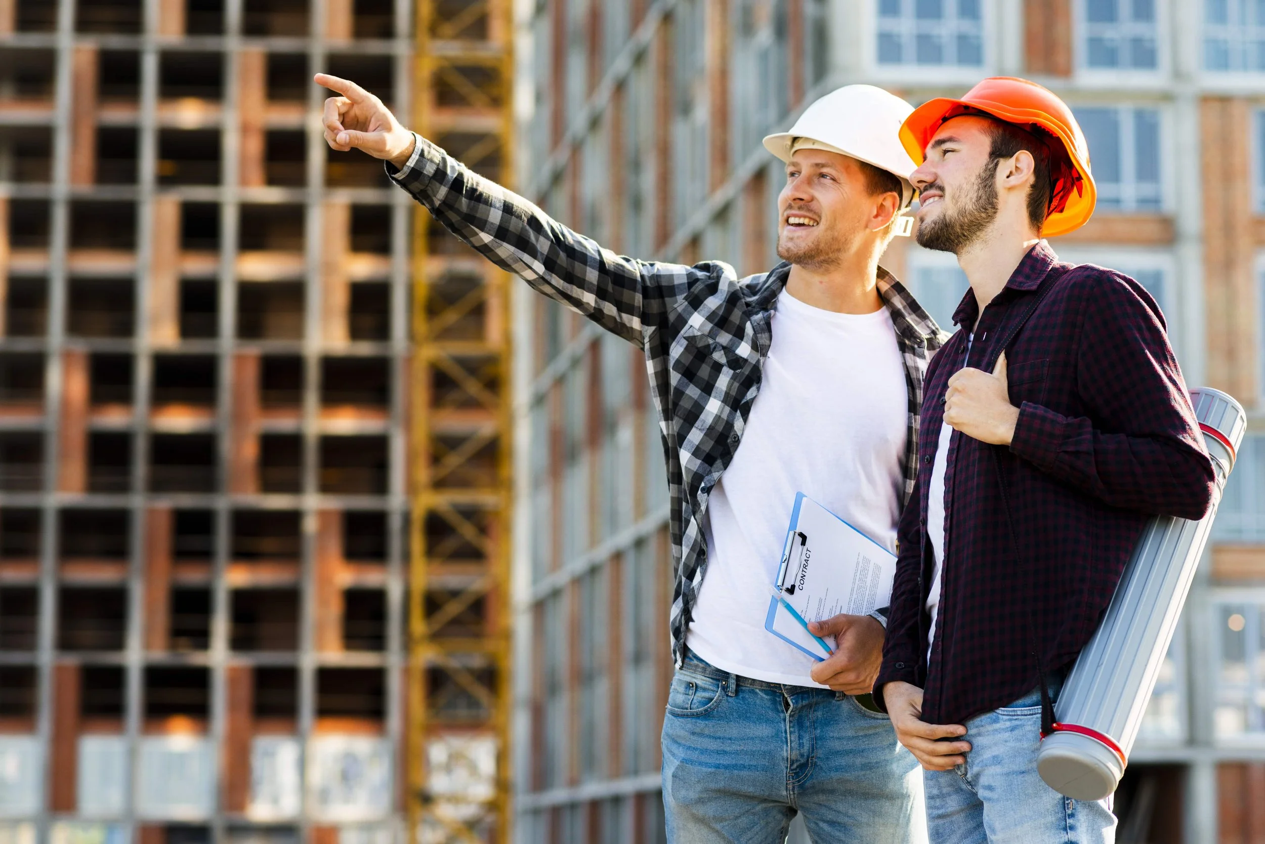 Two construction workers in safety helmets and plaid shirts stand at a building site. One points towards the building, holding a clipboard, while the other carries rolled-up plans.