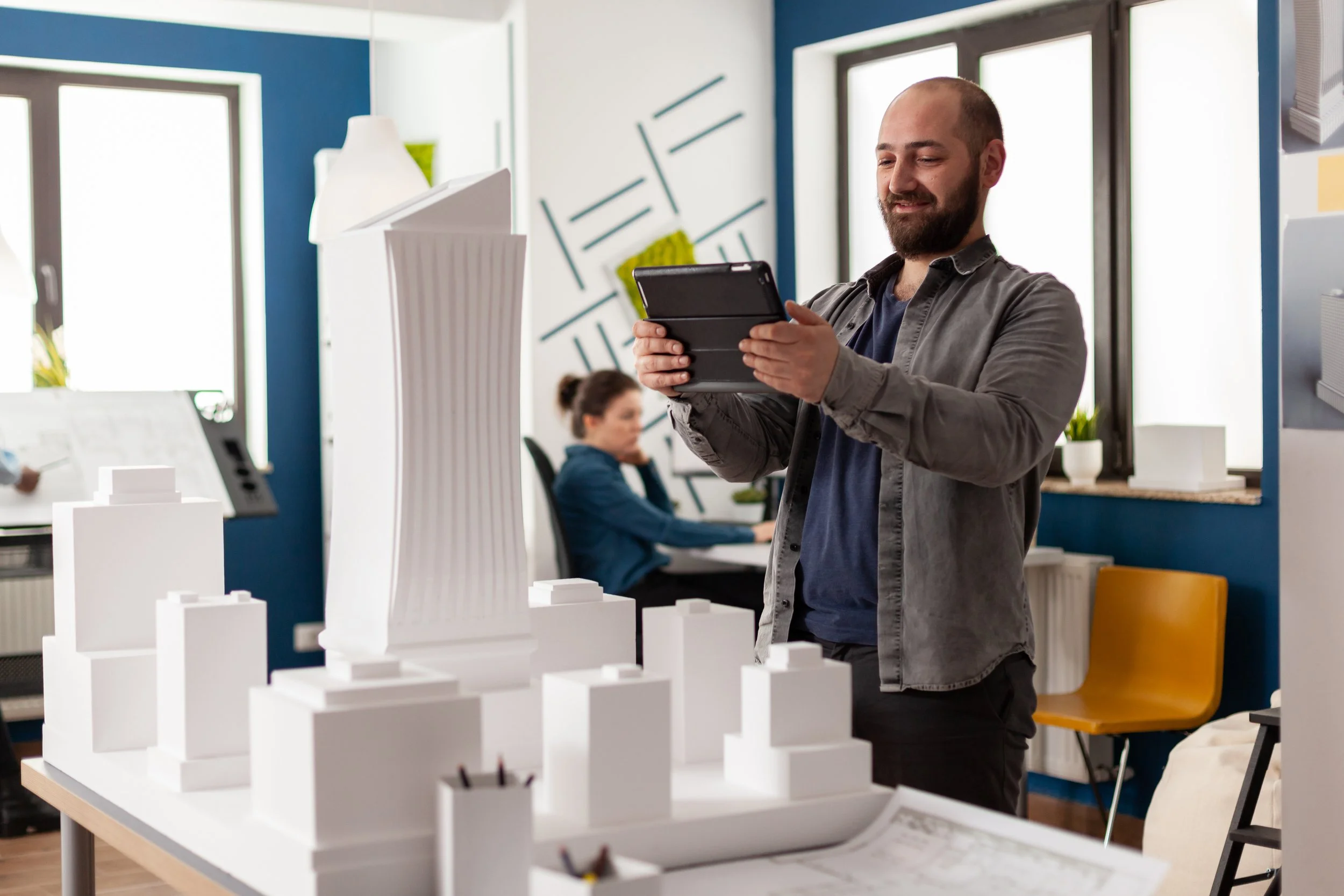 A man in gray shirt and dark pants standing in an office, smiling while holding a tablet, with architectural models on a table in front of him and a woman working at a desk in the background.