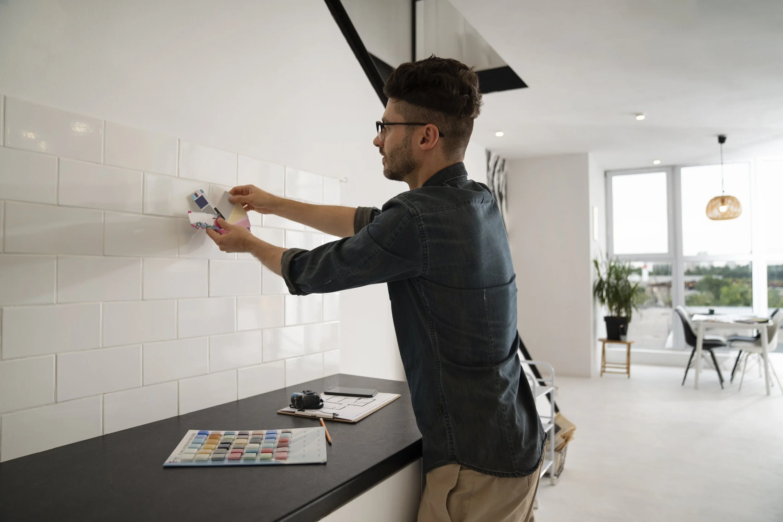 A man in a black denim shirt and glasses is standing at a kitchen counter, applying paint to white tiles with a sponge. On the counter, there are color swatches, a clipboard, and a camera. A living room with large windows, a dining table, chairs, and a potted plant is visible in the background.