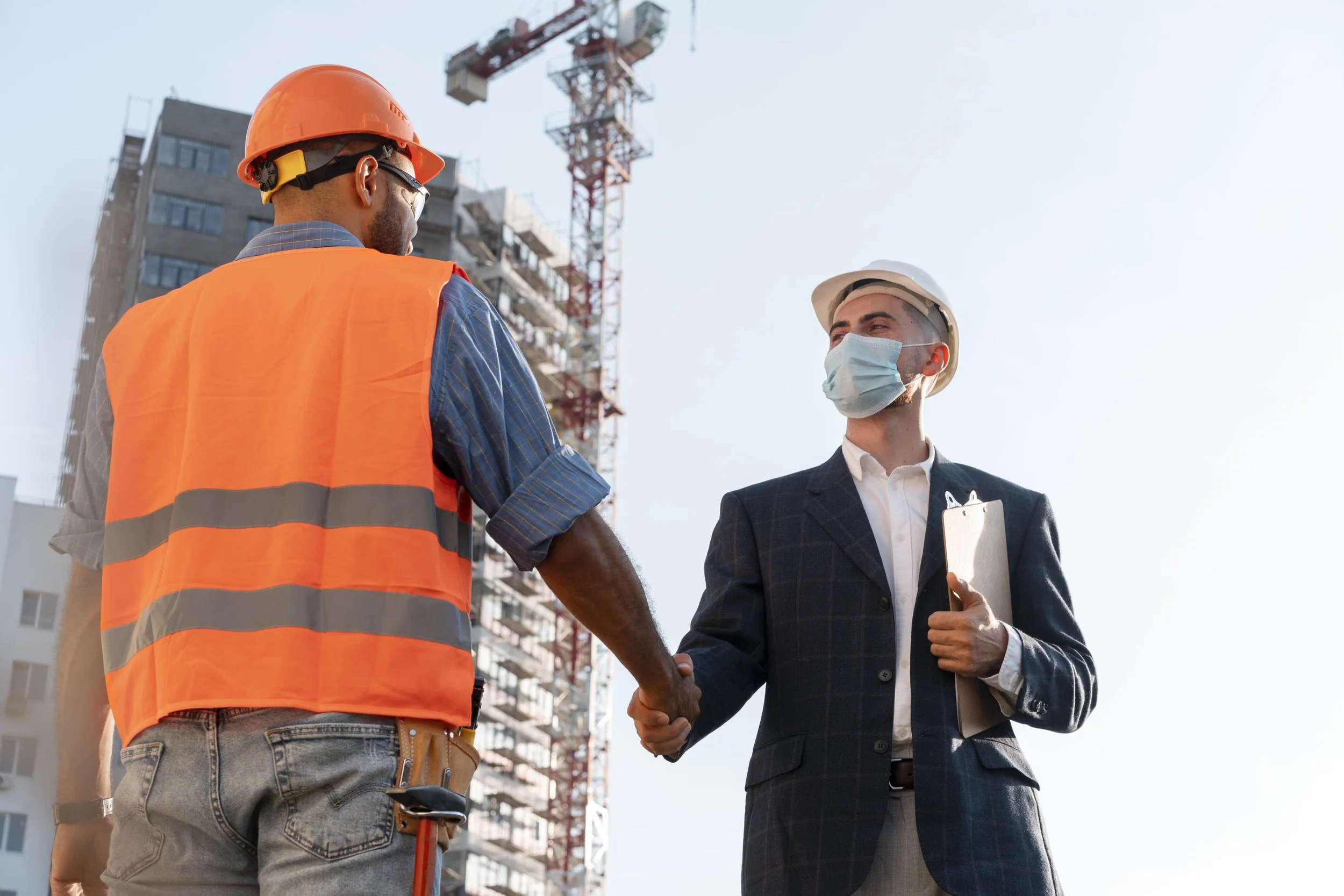 Two men in safety helmets and masks shaking hands on a construction site with a building and crane in the background.