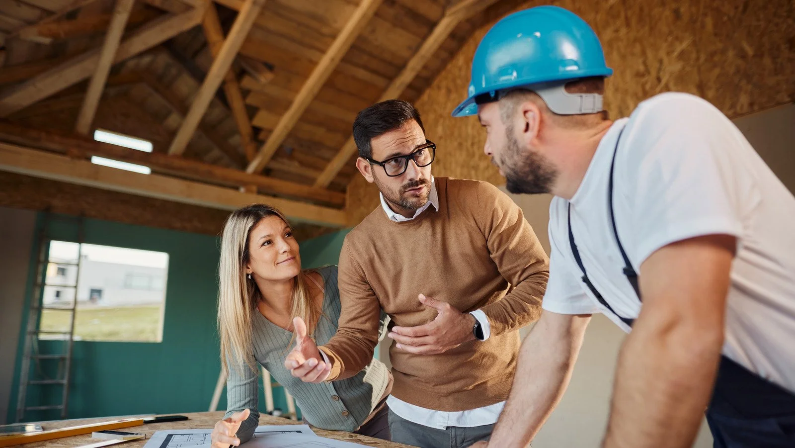 Three people having a discussion inside a house under construction, with one person wearing a construction helmet.