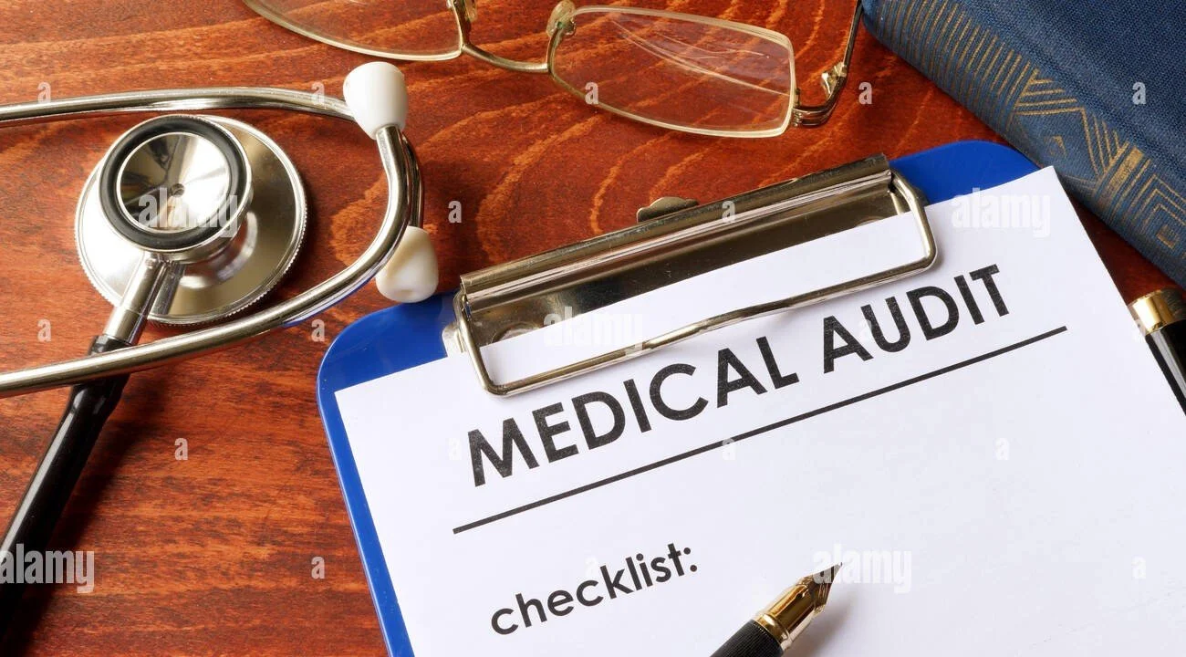 A clipboard with a medical audit checklist, a pen, a stethoscope, eyeglasses, and a blue book on a wooden desk.