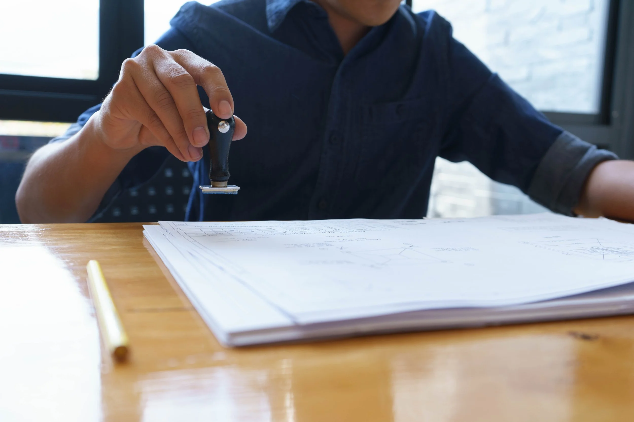 Person holding a stamp over architectural blueprints on a wooden table, with a yellow pencil to the left.