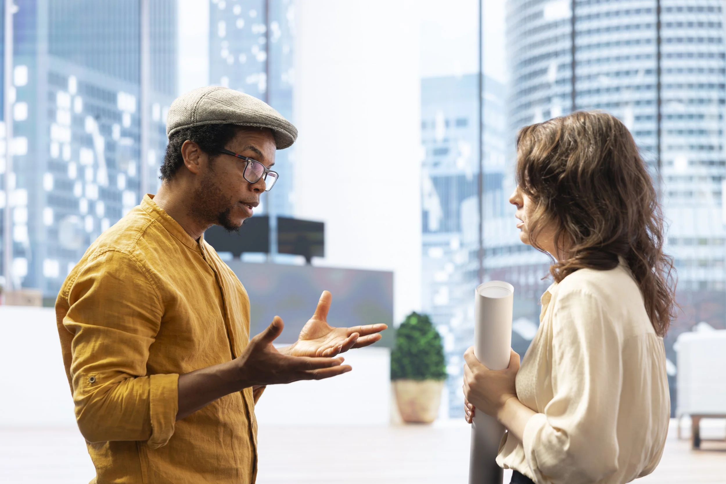 Two people having a conversation in an office with large windows and cityscape background, one man wearing a yellow shirt and gray hat, the woman wearing a beige blouse and holding rolled paper.