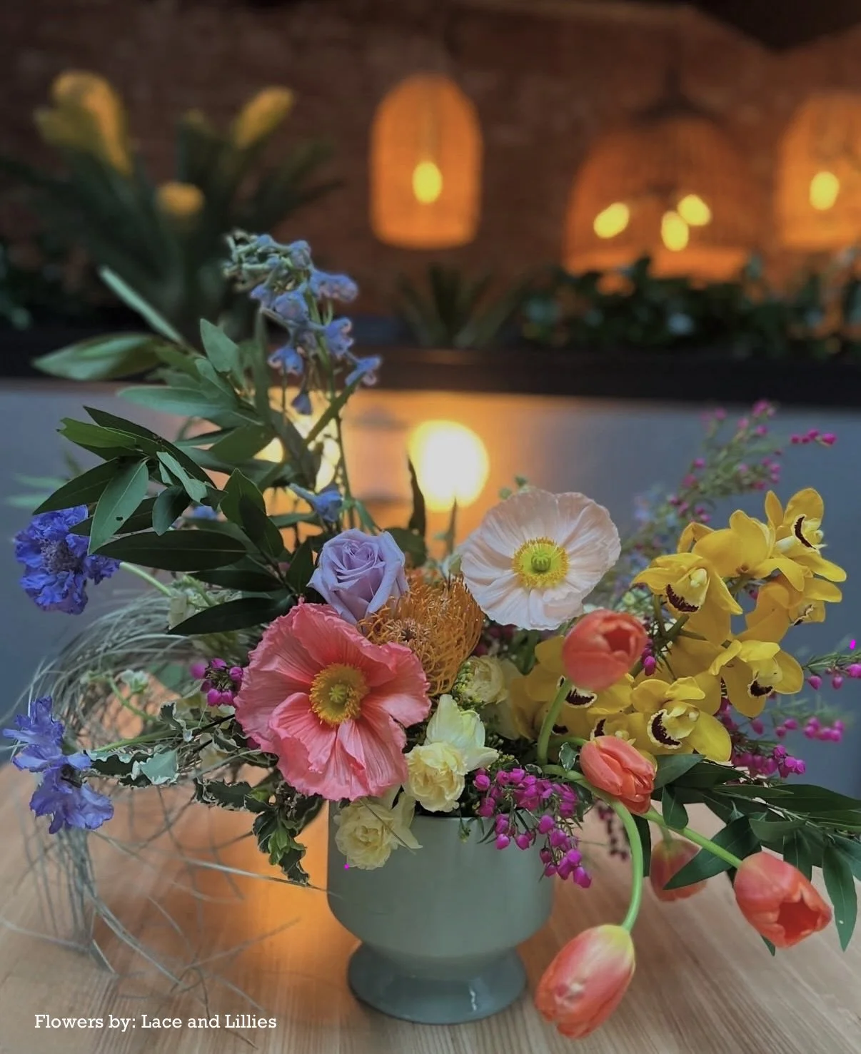 Colorful wedding bouquet of flowers including pink poppies, yellow orchids, purple roses, and blue blooms in a gray vase on a wooden table with a candle reflection, with warm hanging lights in the background.
