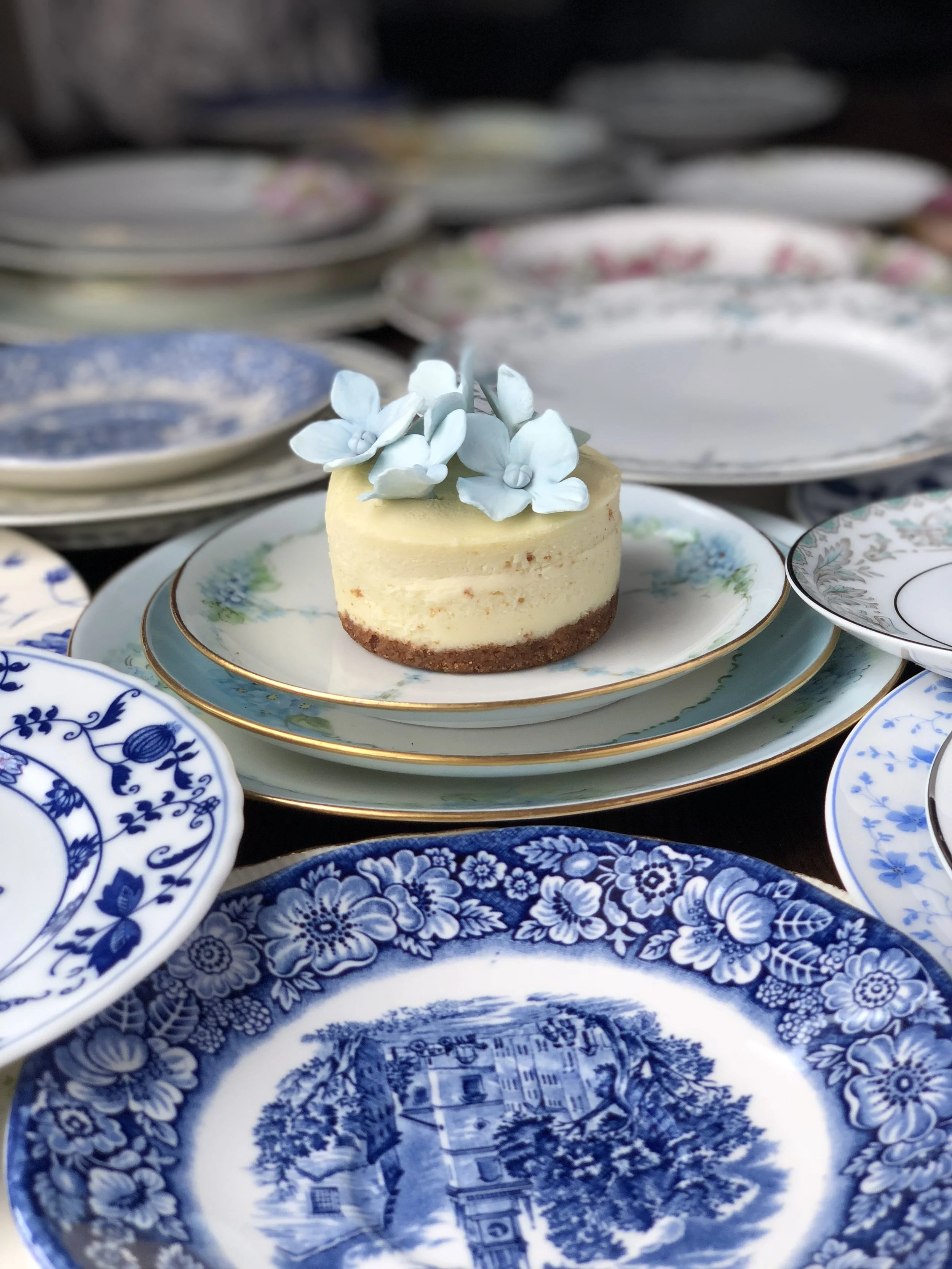 A small round cheesecake with white flowers on top, placed on a decorative floral plate among an array of blue and white patterned porcelain plates at a wedding.