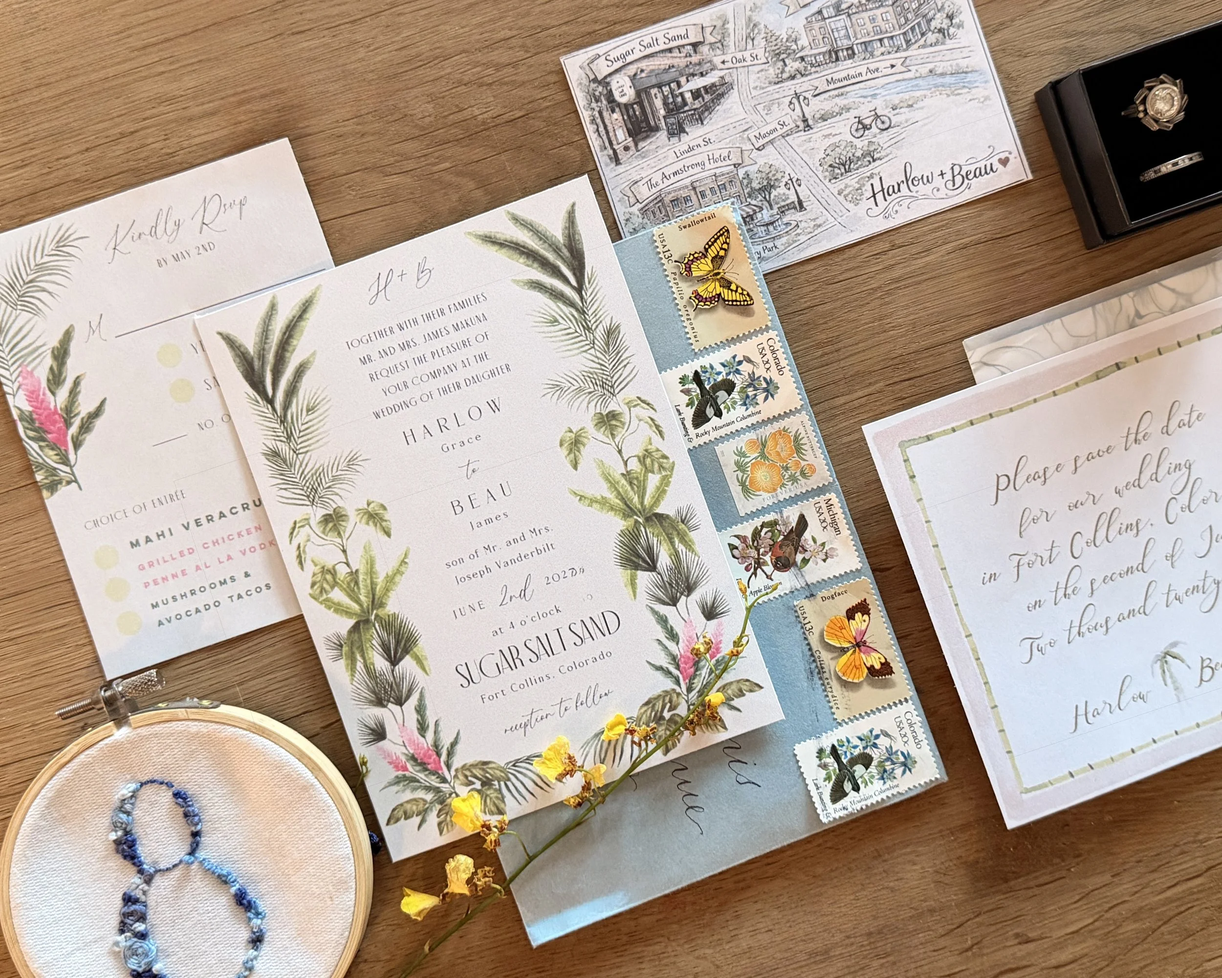 Wedding invitation with botanical illustrations, postage stamps, a save-the-date card, a ring in a black box, and a small embroidered table number, all arranged on a wooden surface.