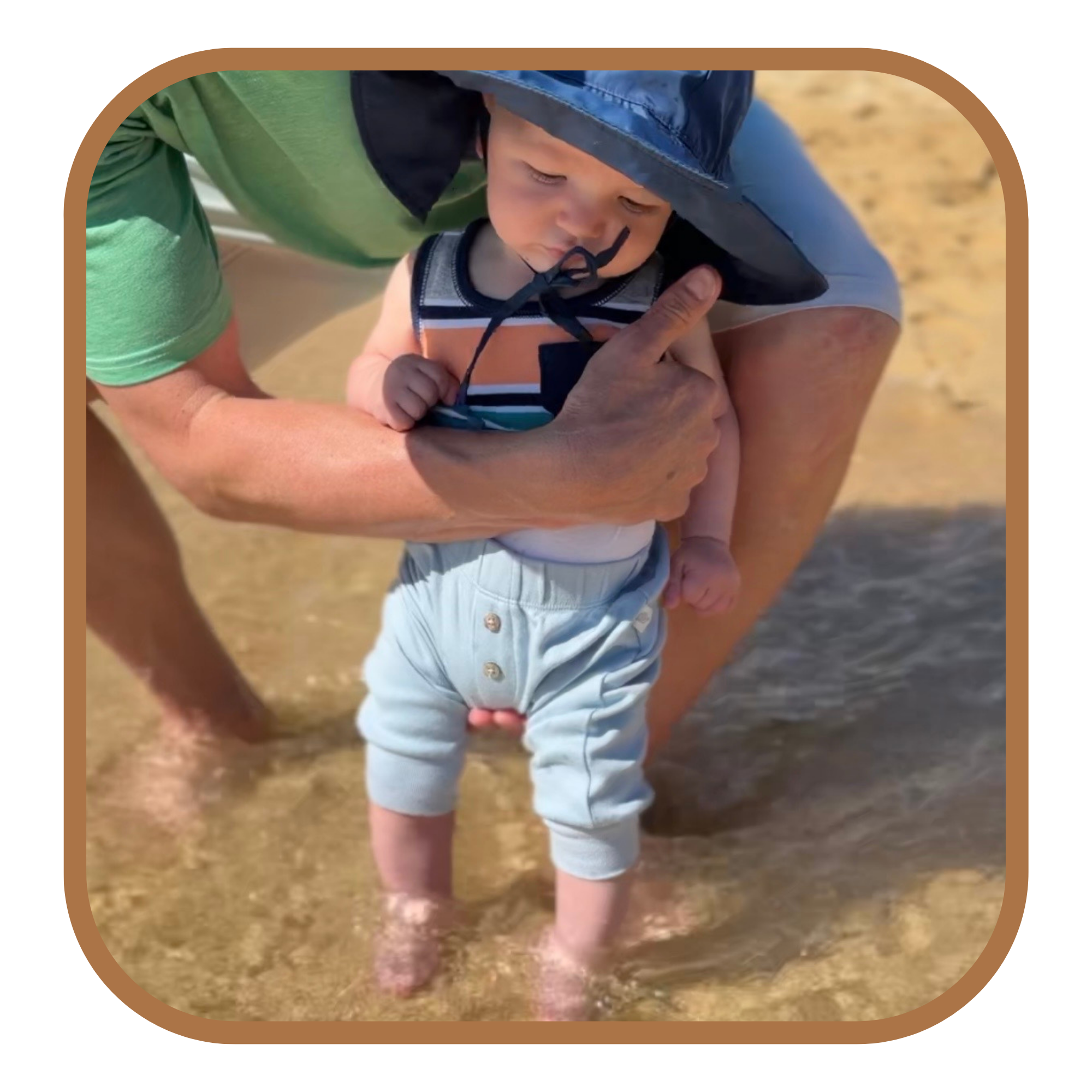 A young child standing in shallow water at the beach, supported by an adult's arms. The child is wearing a striped tank top, light blue pants, and a sun hat. The adult is kneeling behind the child, holding him securely, and is wearing a green shirt and shorts.