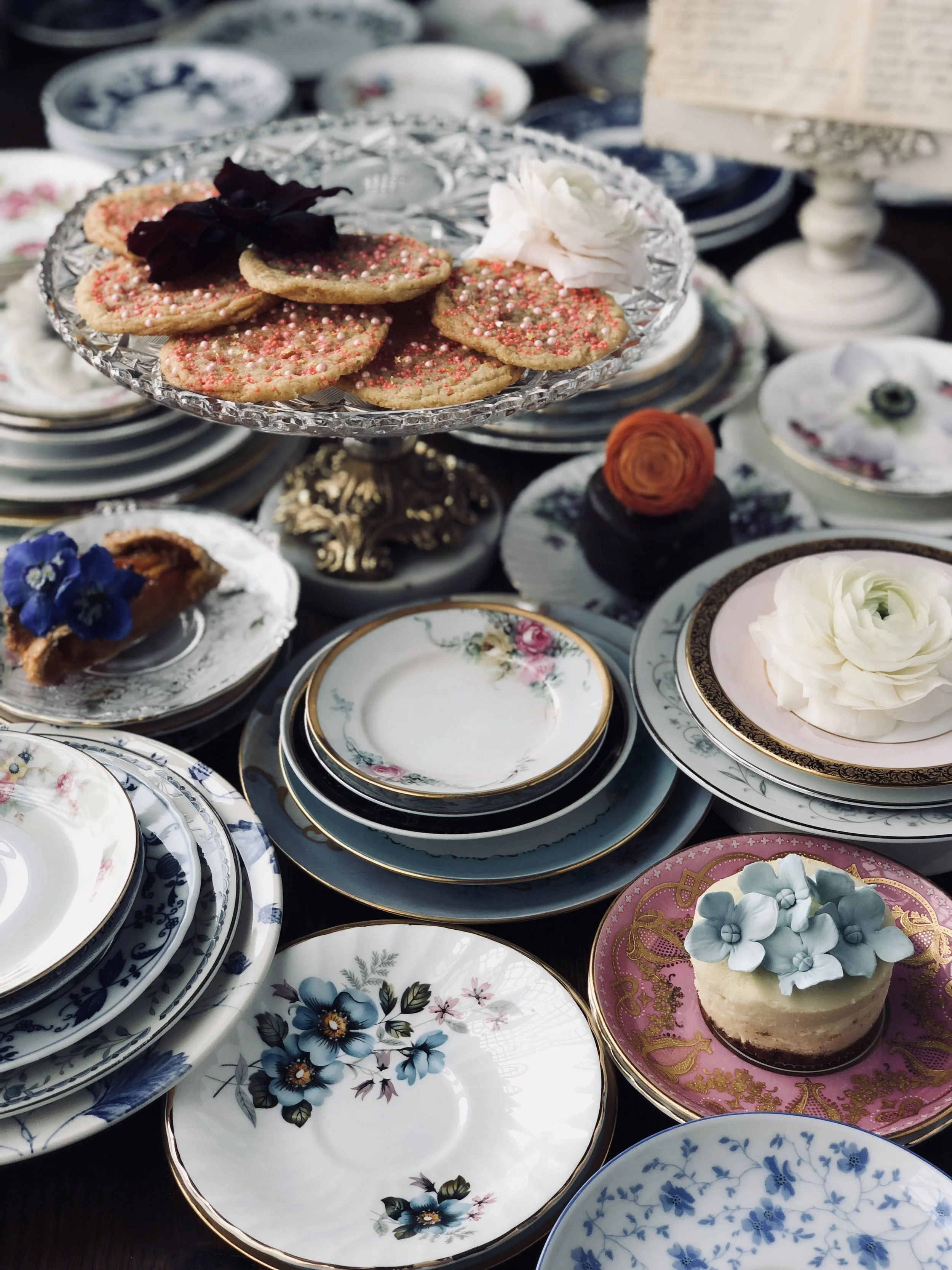 An assortment of vintage floral patterned china plates and cups, with a glass cake stand holding decorated cookies topped with sprinkles and flowers, surrounded by an array of decorative desserts and pastries.