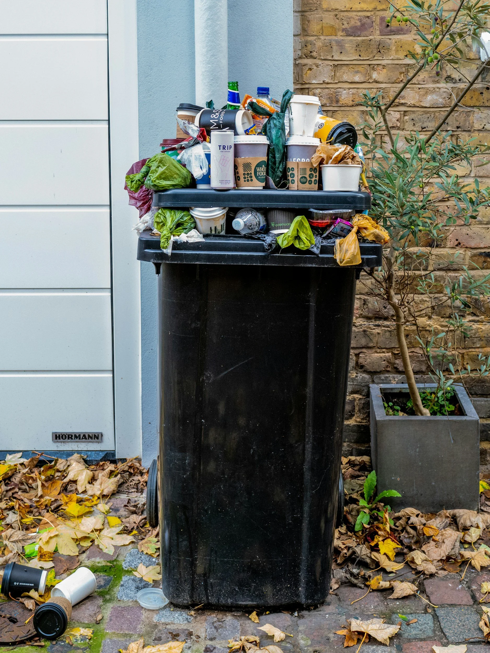 A black garbage bin overfilled with food waste and other garbage in front of residential garage door