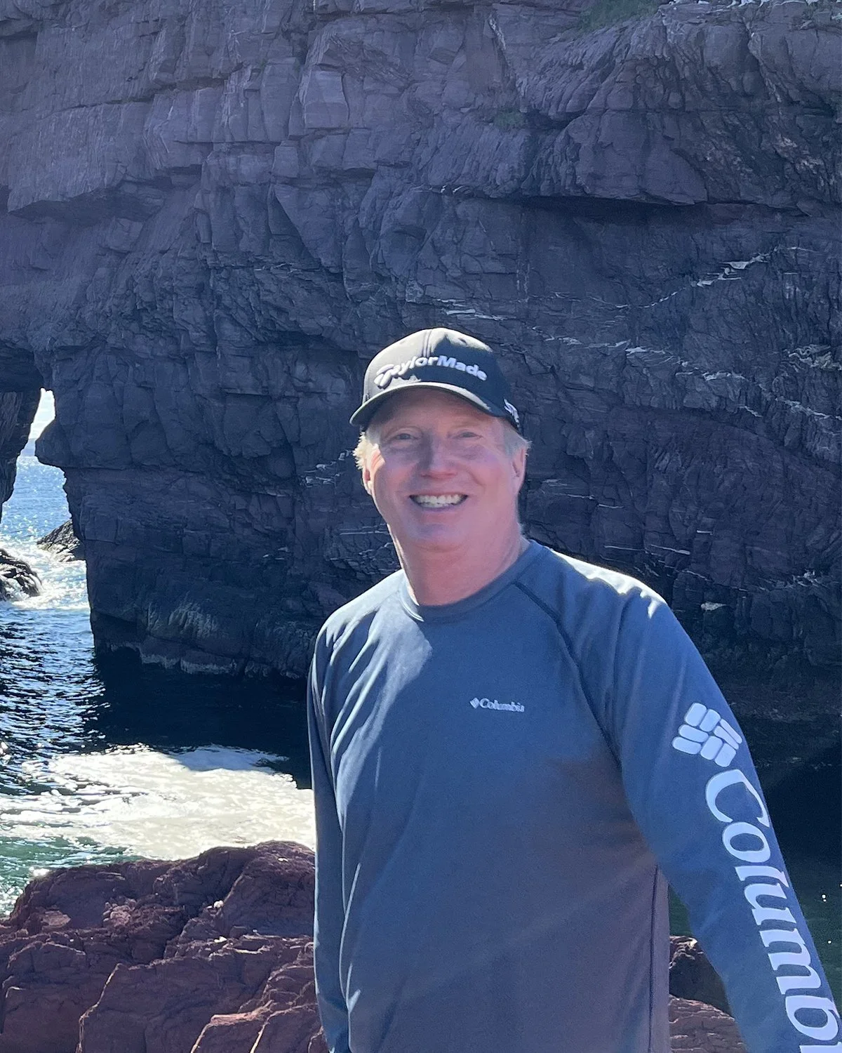 Headshot of JOHN MACRAE, ecoCaledon co-chair, smiling outside on a beach with rocks behind him