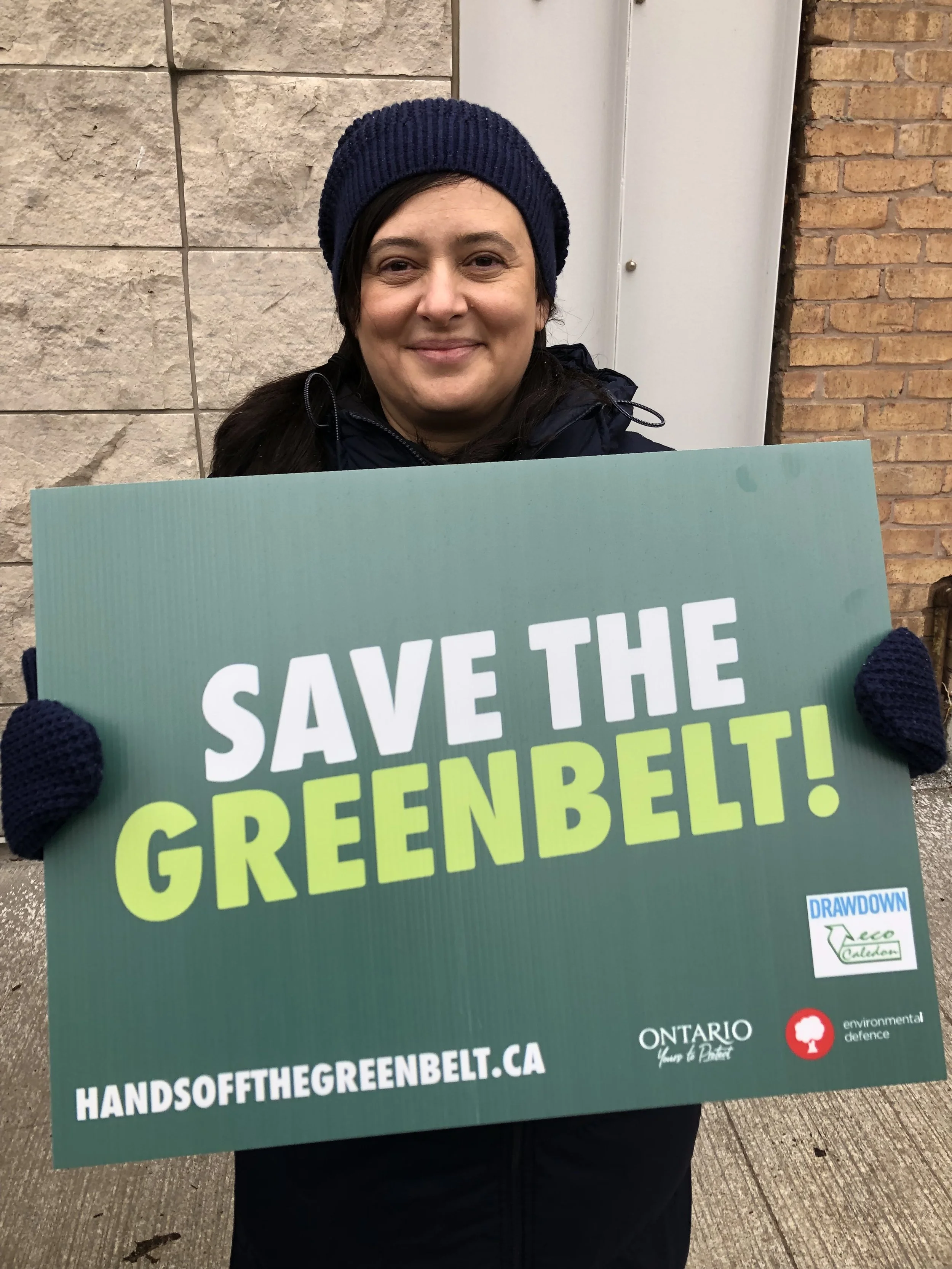 Headshot of LUCREZIA CHIAPPETTA, ecoCaledon co-chair, smiling and holding a "Save the Greenbelt" sign outside at an event
