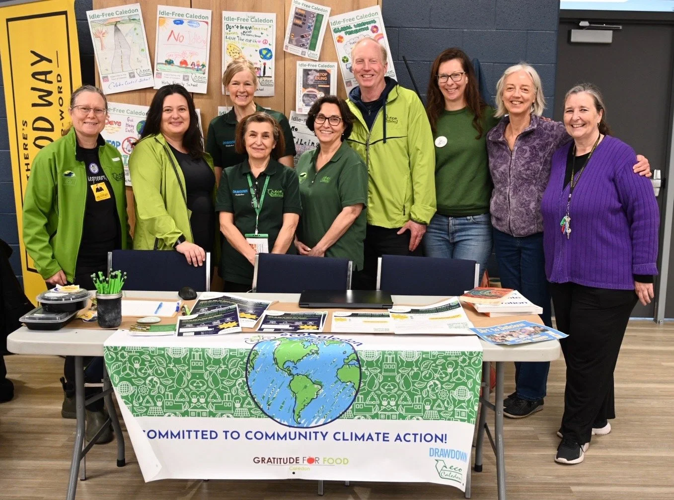 ecoCaledon members standing and smiling together, in front of fold out table with banner on it that says "Committed to Community Climate Action" Gratitude For Food ecoCaledon
