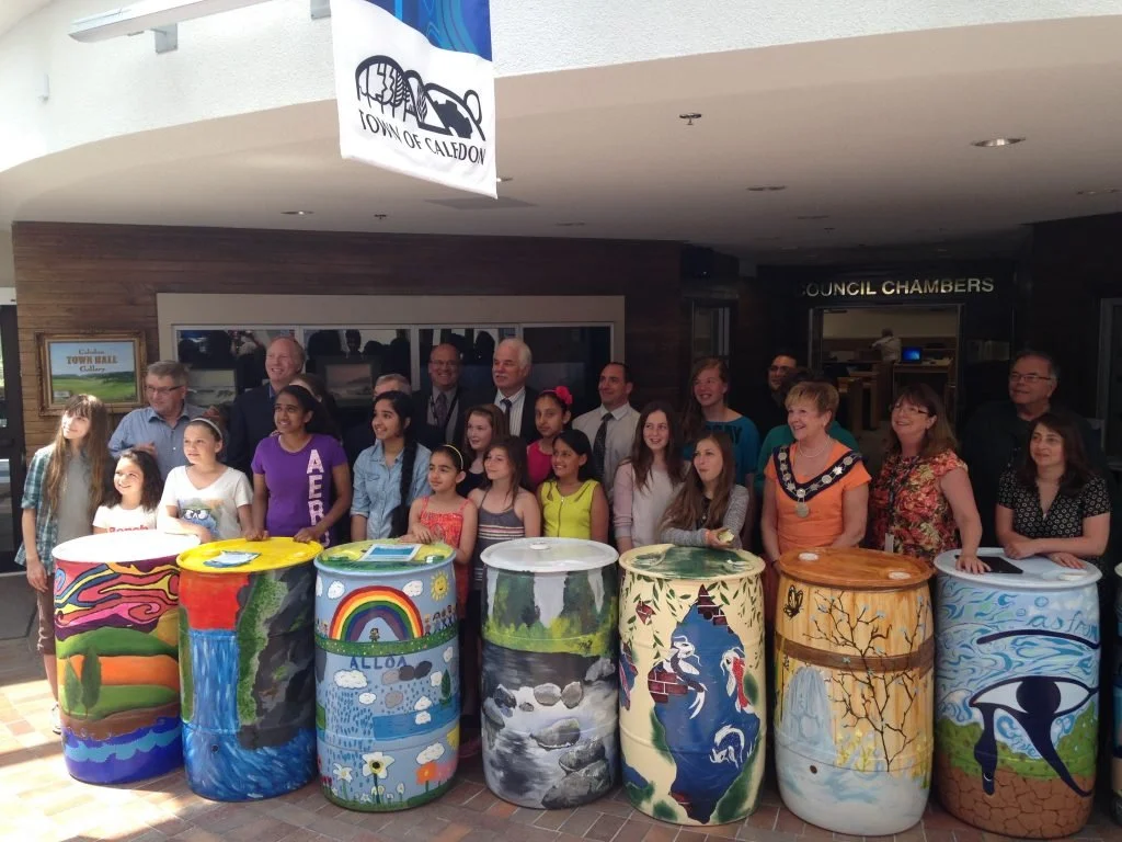 Caledon-based kids and adult community members smiling in a group with painted rain barrels in front of the Town of Caledon Council Chambers, from an ecoCaledon rain barrel project