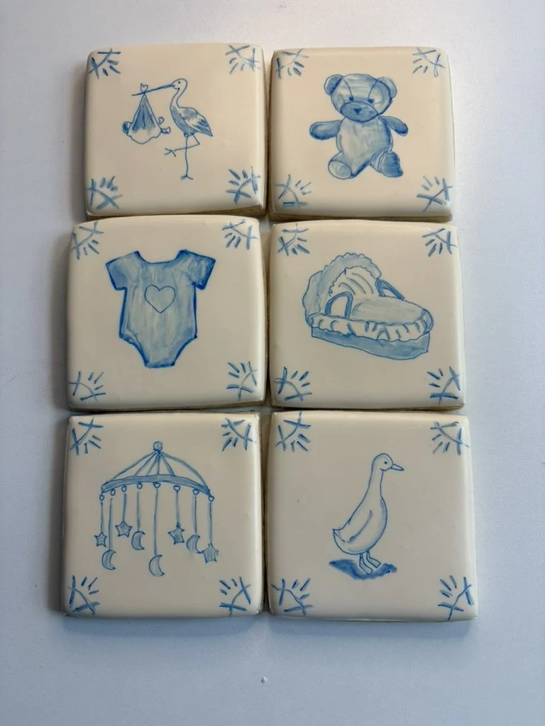 Six white tiles with blue drawings. Top row: a stork carrying a baby blanket, a teddy bear. Middle row: a baby onesie with a heart, a dinosaur skull. Bottom row: a baby mobile with moons and stars, a baby duckling.