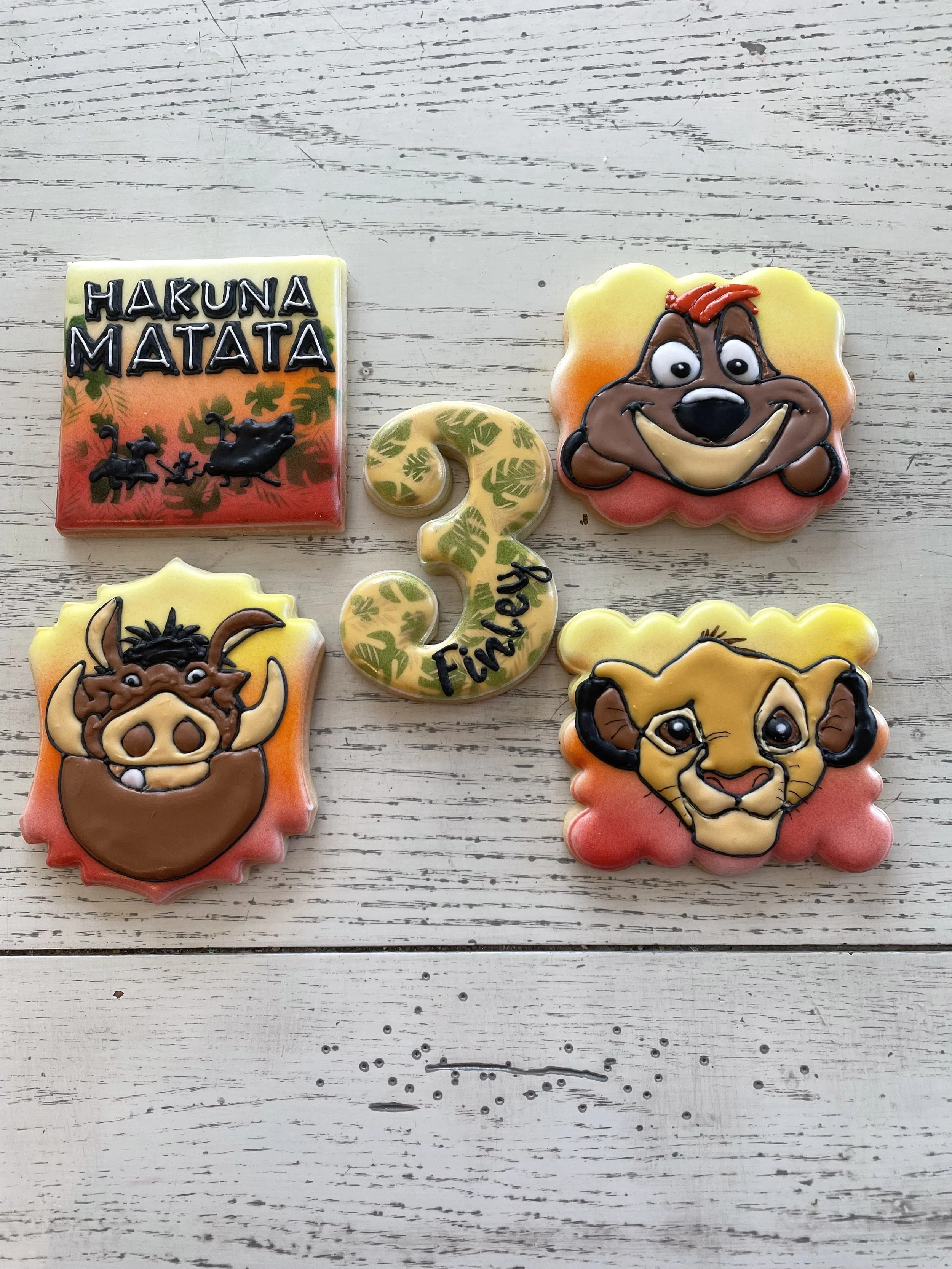 Colorful themed cookies with Lion King characters and a number three cookie that says 'Birthday', arranged on a white wooden surface.