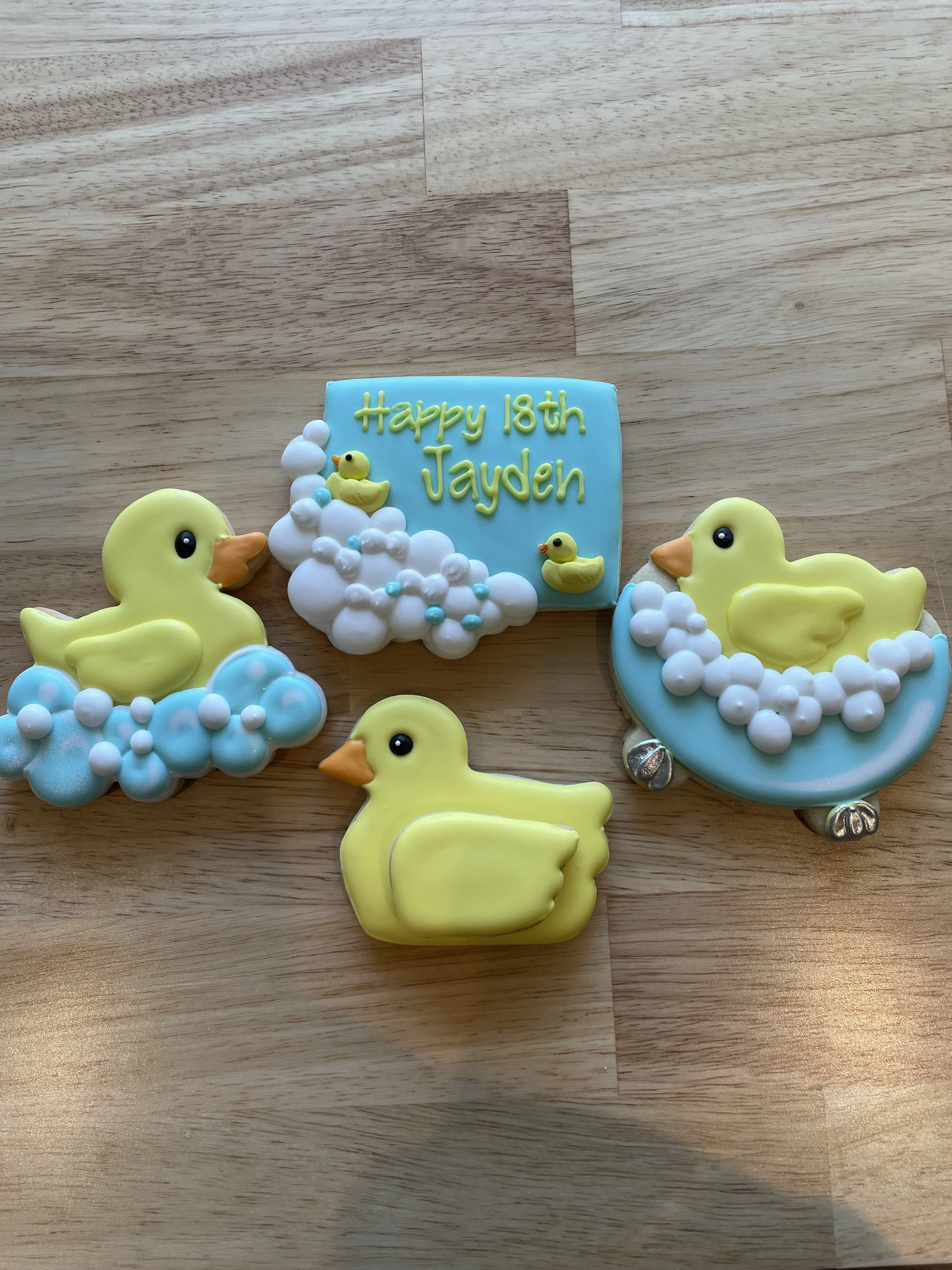 Decorative cookies with yellow rubber duck designs, clouds, and a blue message card saying "Happy 18th Jayden," on a wooden surface.