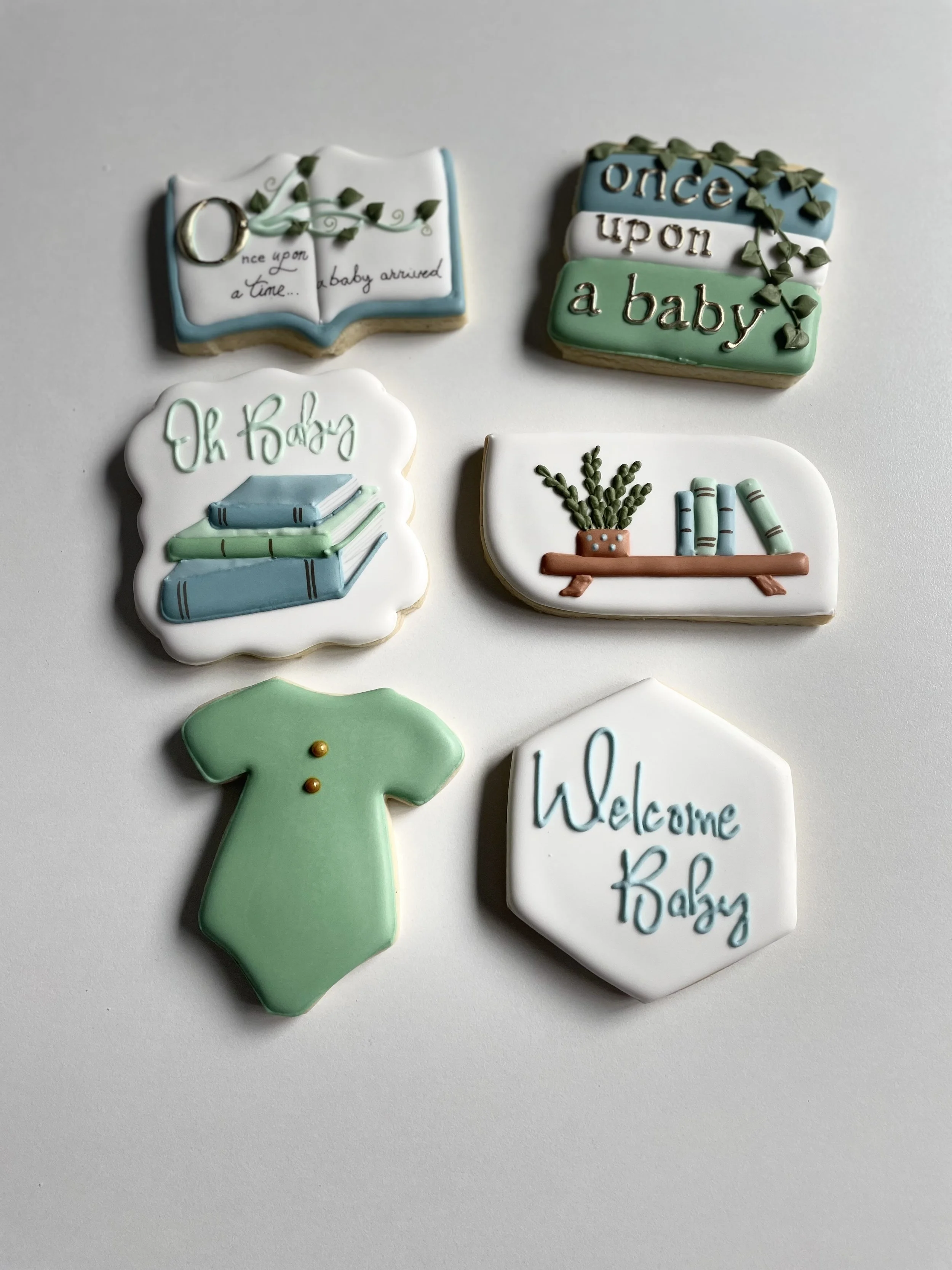 Set of decorated cookies celebrating the arrival of a baby, including an open book with the words "Once upon a baby arrived," a sign with "once upon a baby," a cloud shaped cookie with "Oh Baby" and stacked books, a bookshelf with books and a plant,