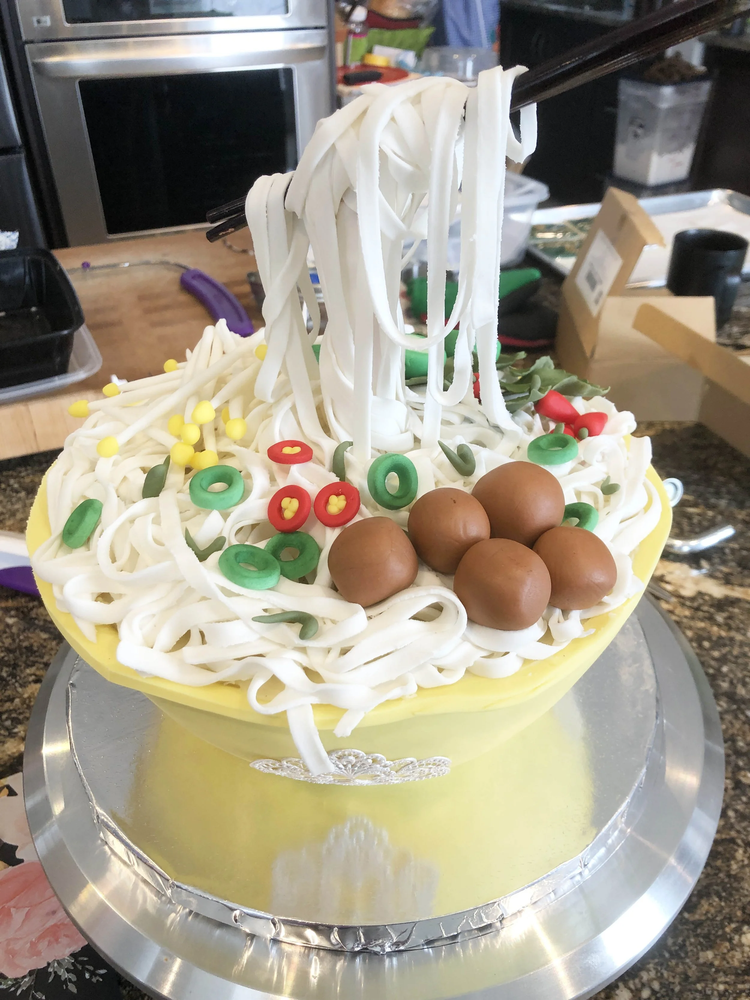 A cake decorated to look like a bowl of noodles with brown meatballs, colorful candy toppings, and chopsticks.