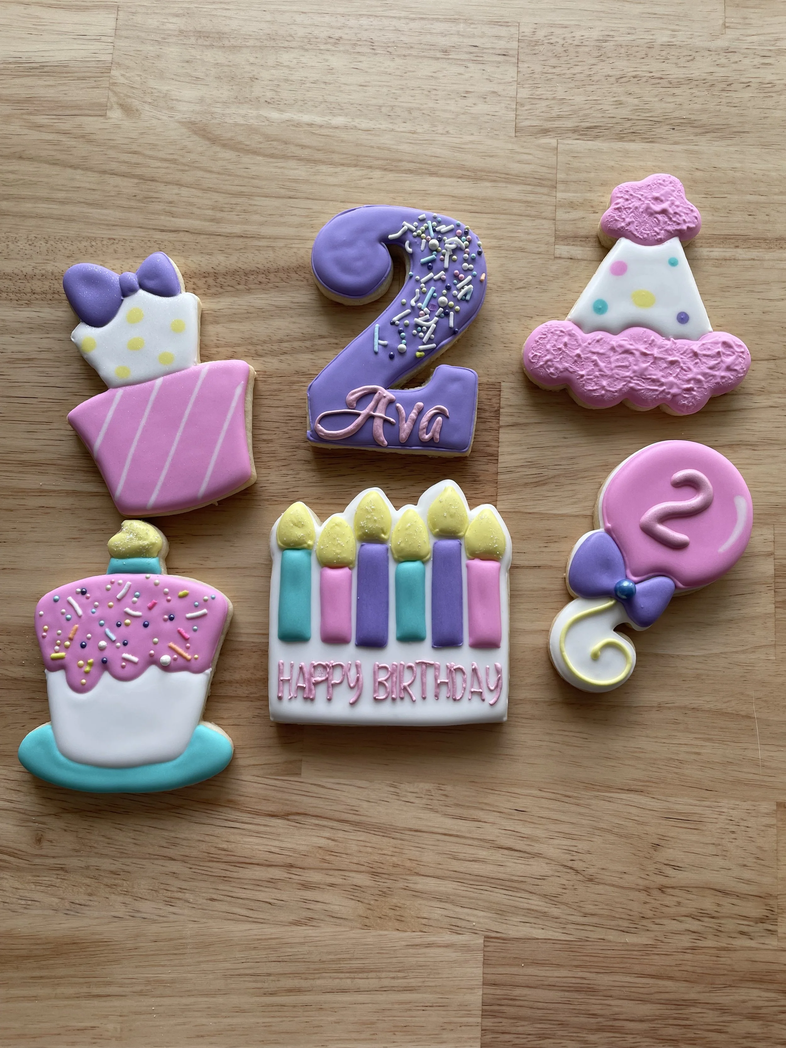 Decorative birthday cookies on a wooden surface, including a cake slice with a bow, a purple number 2 with sprinkles and the name 'Ava', a birthday hat, a decorated candle cake, a balloon with a number 2 and bow, and a birthday cake with candles and