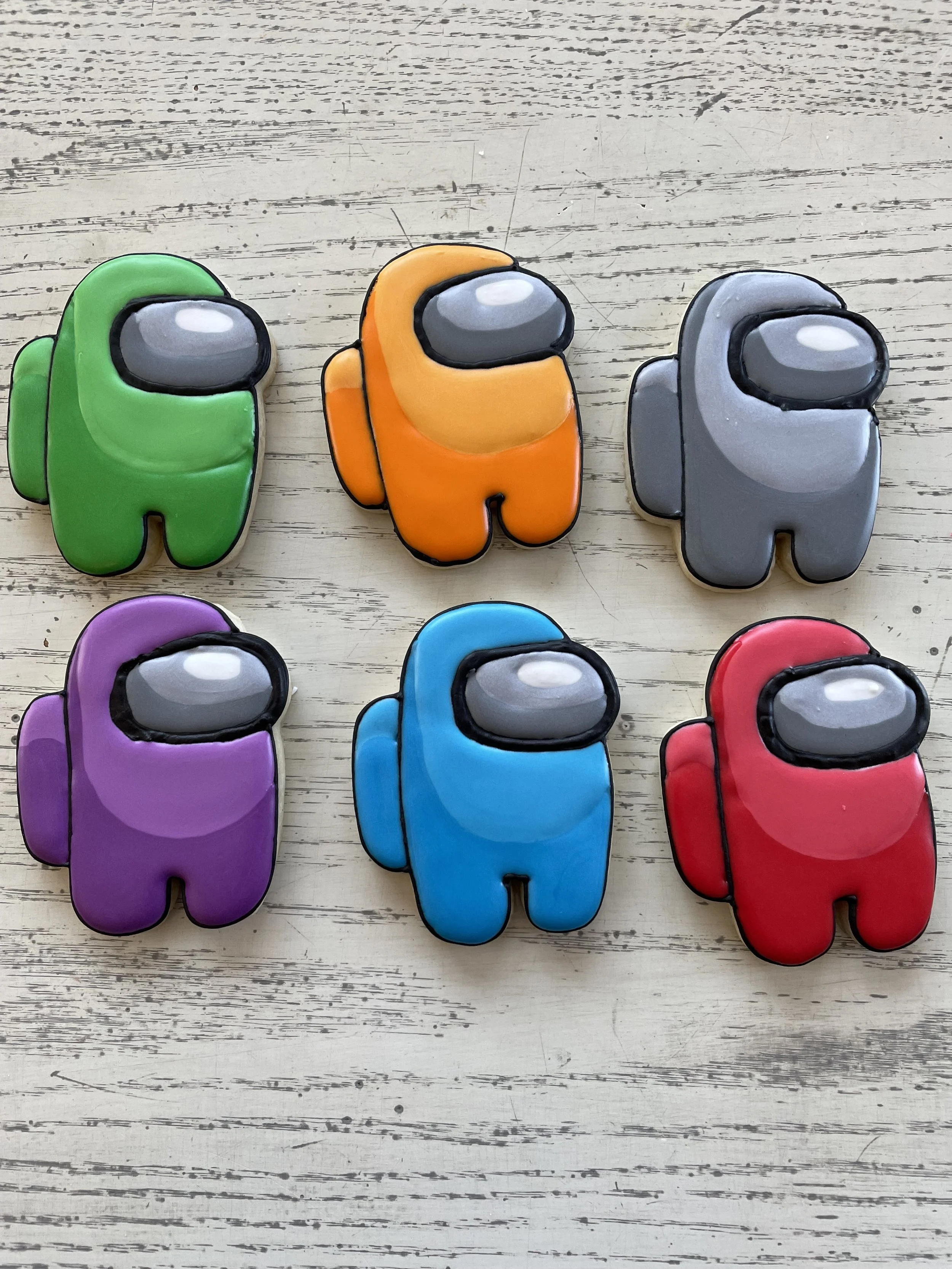 Colorful cookies shaped like characters from Among Us, with green, orange, gray, purple, blue, and red colors, on a wooden surface.
