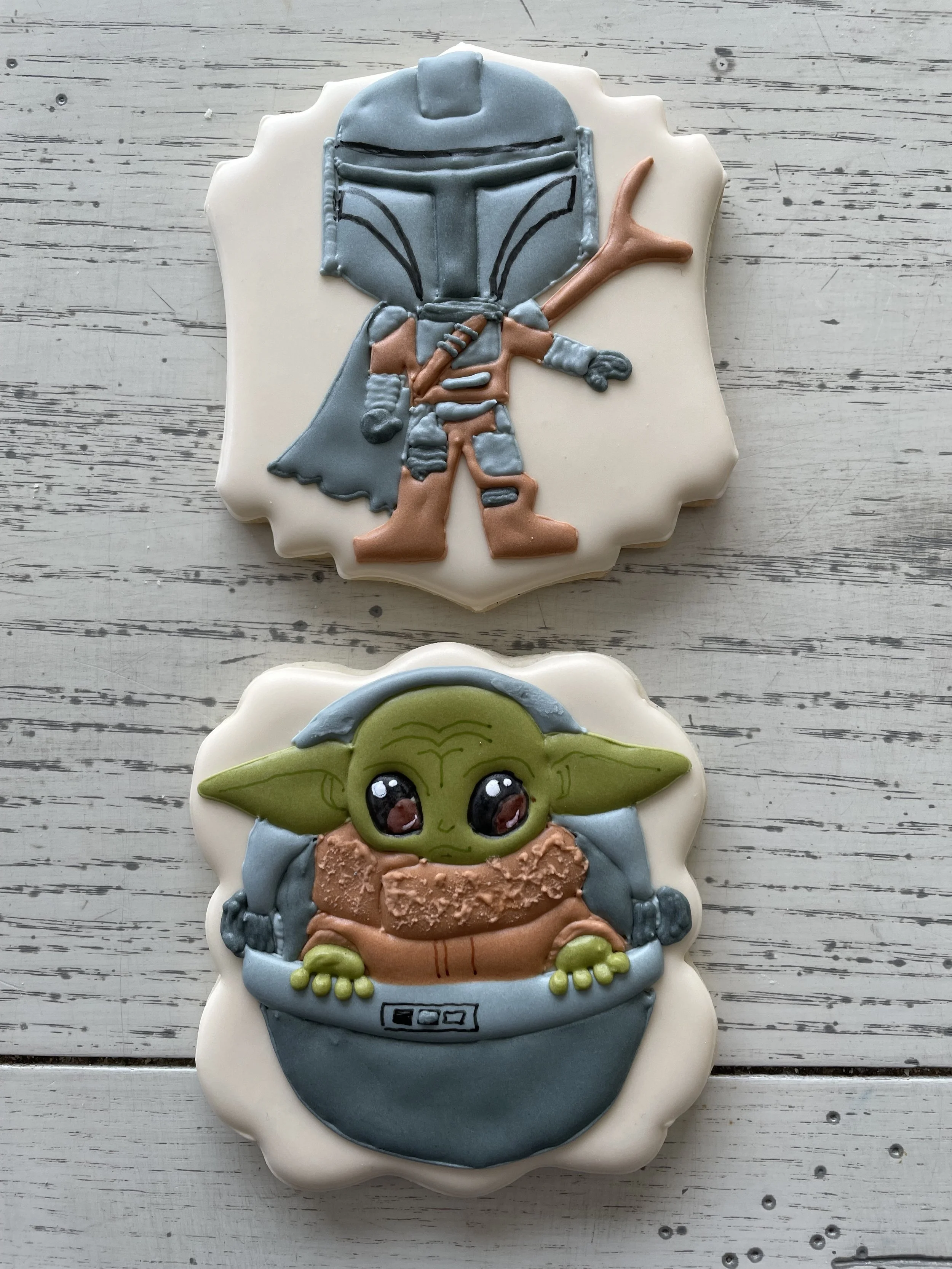 Star Wars themed cookies featuring a Mandalorian with a helmet and armor, and Baby Yoda (Grogu) with large eyes and green skin, both decorated with detailed icing.