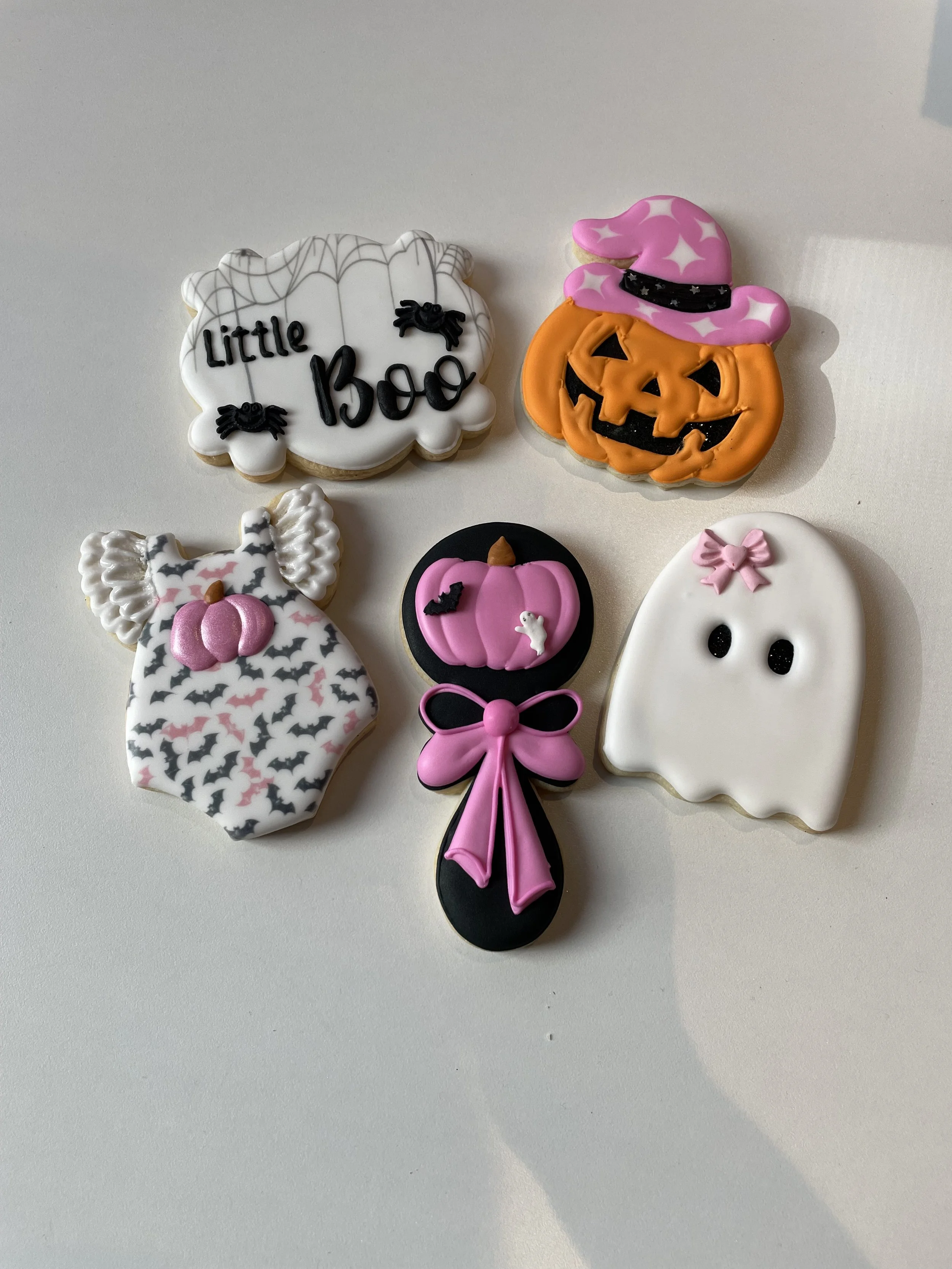 Collection of five decorated Halloween cookies on a white surface, including a ghost, a pumpkin with a witch hat, a bat on a pink pumpkin, a white ghost with pink bow, and a cookie with the text 'Little Boo' with spider decorations.