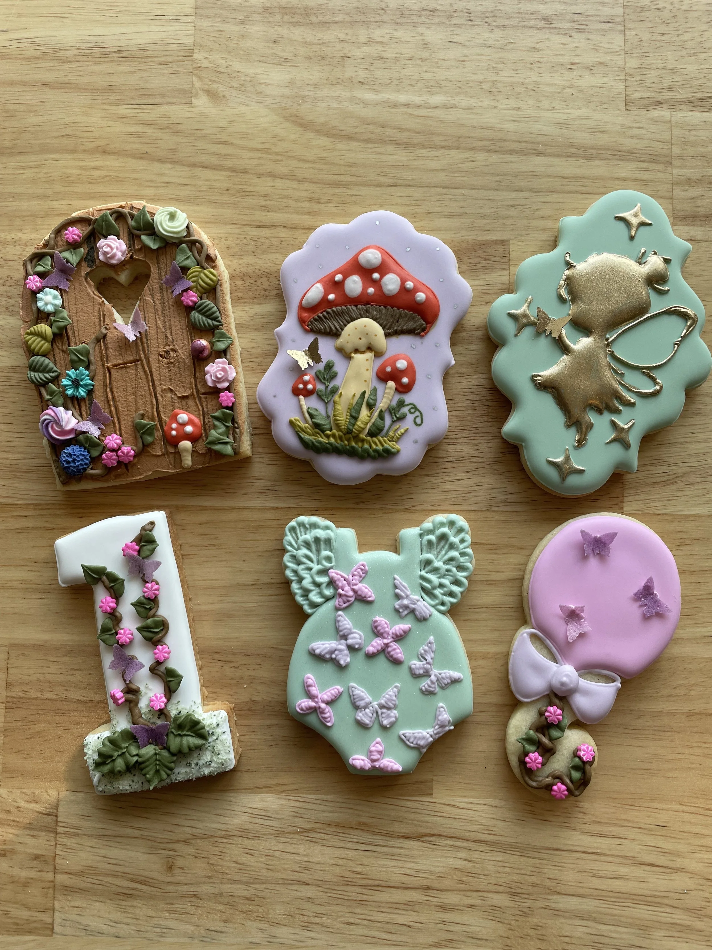 Colorful decorated cookies shaped like the number one, mushroom, fairy, butterfly, and balloon with flowers, on a wooden surface.