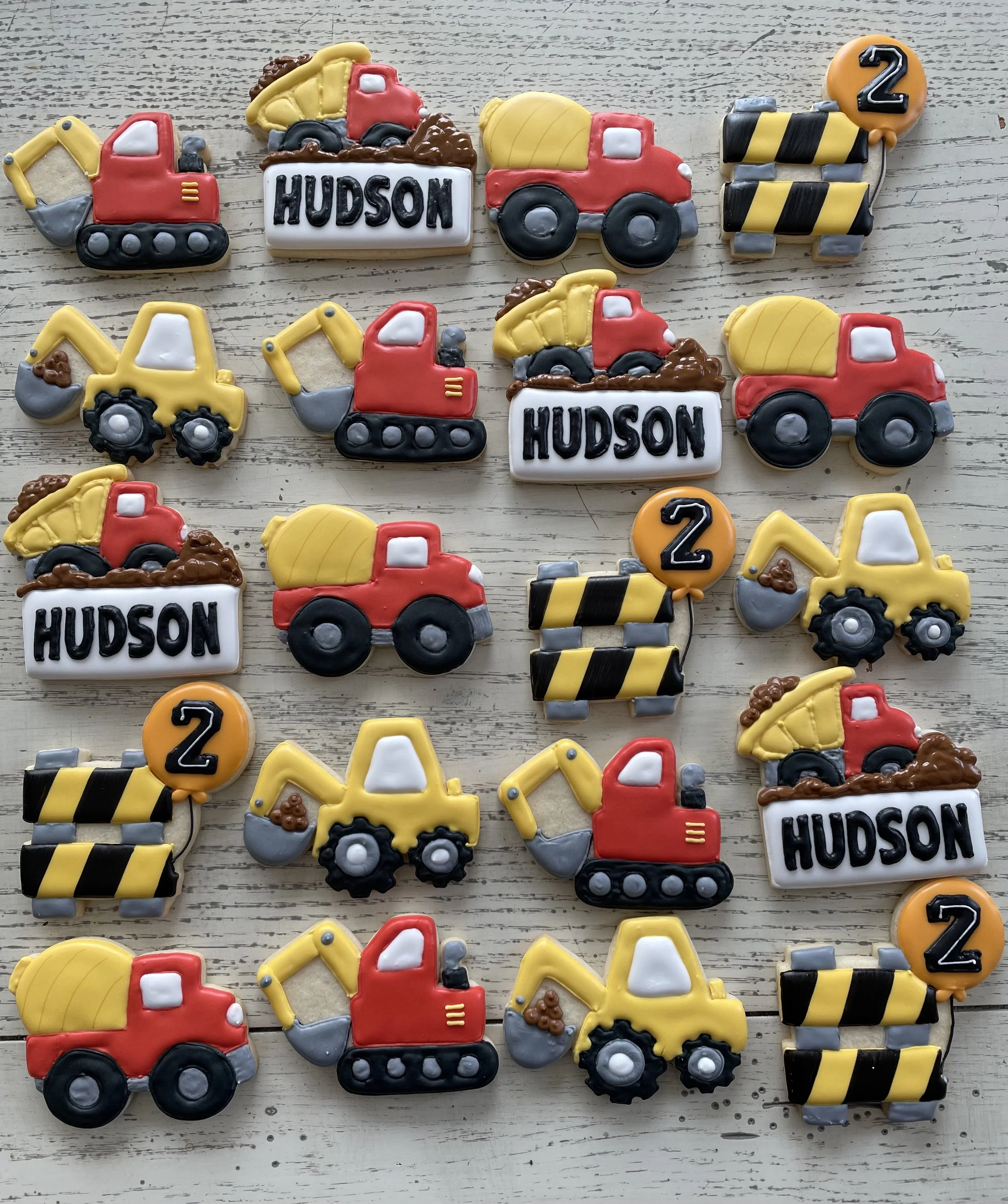 Cookies decorated as construction trucks, with words "Hudson" and numbers "2" on some cookies, arranged on a wooden surface.
