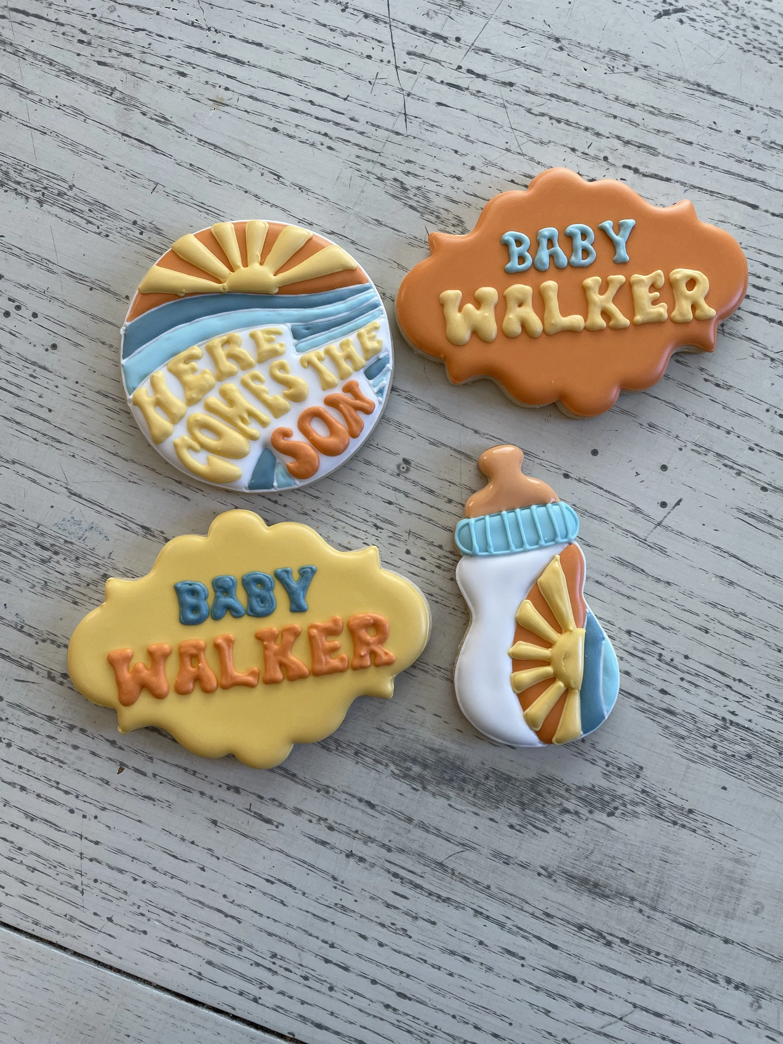 Decorative cookies themed for a baby's walk, with colorful icing and text reading 'Here Comes the Son' and 'Baby Walker', placed on a white wooden surface.