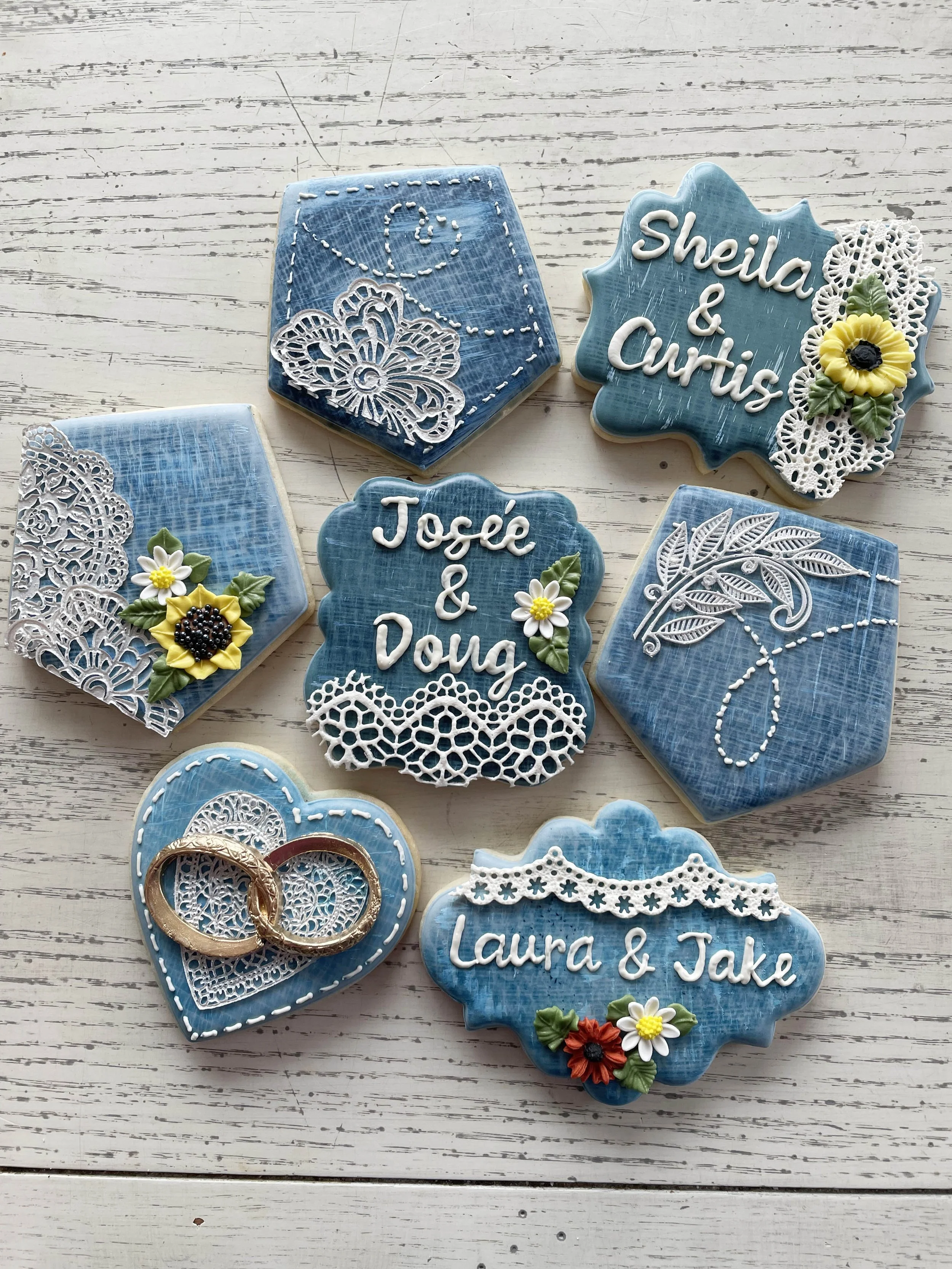 Decorated sugar cookies with blue icing, floral decorations, and wedding-related text, including names Sheila & Curtis, Josie & Doug, and Laura & Jake, arranged on a white wooden surface.