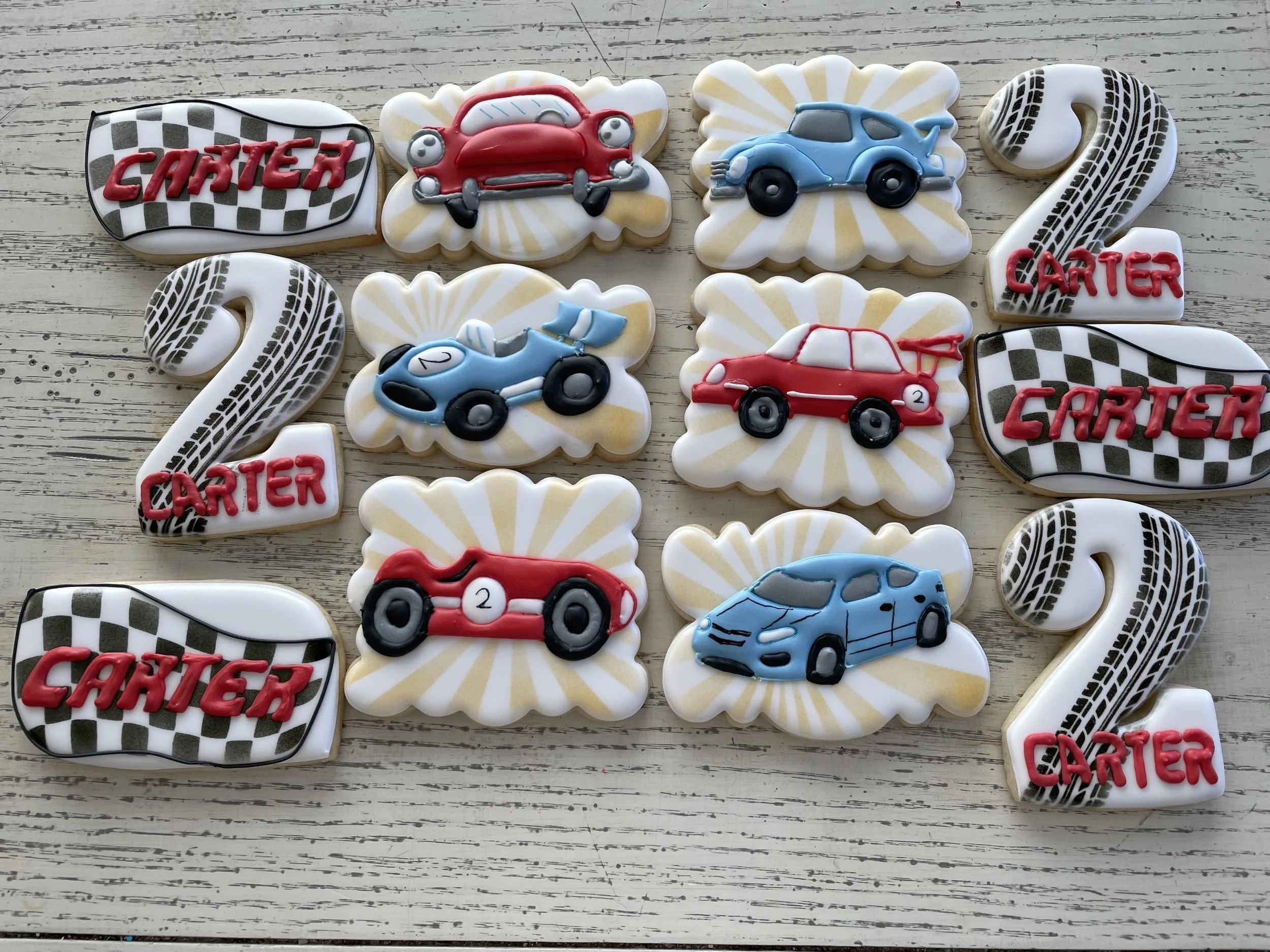 Cookies decorated with race car themes including race cars, race flags, and the number 2, arranged on a white wooden surface.