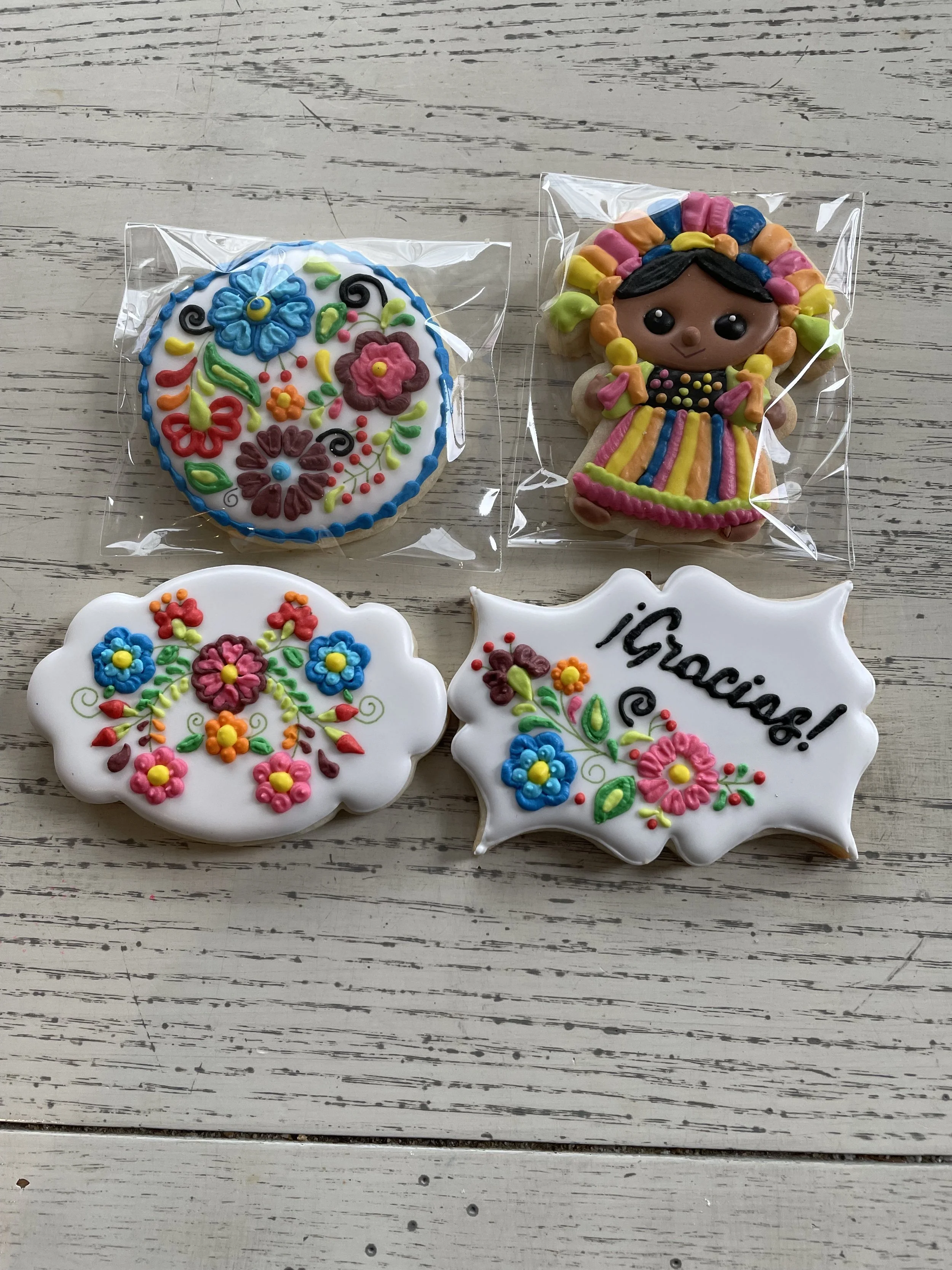 Decorative cookies on a white wooden surface, including a round cookie with colorful floral icing, a figure cookie of a girl with a rainbow-colored dress and hair, a cloud-shaped cookie with floral icing, and a white oval-shaped cookie with the word
