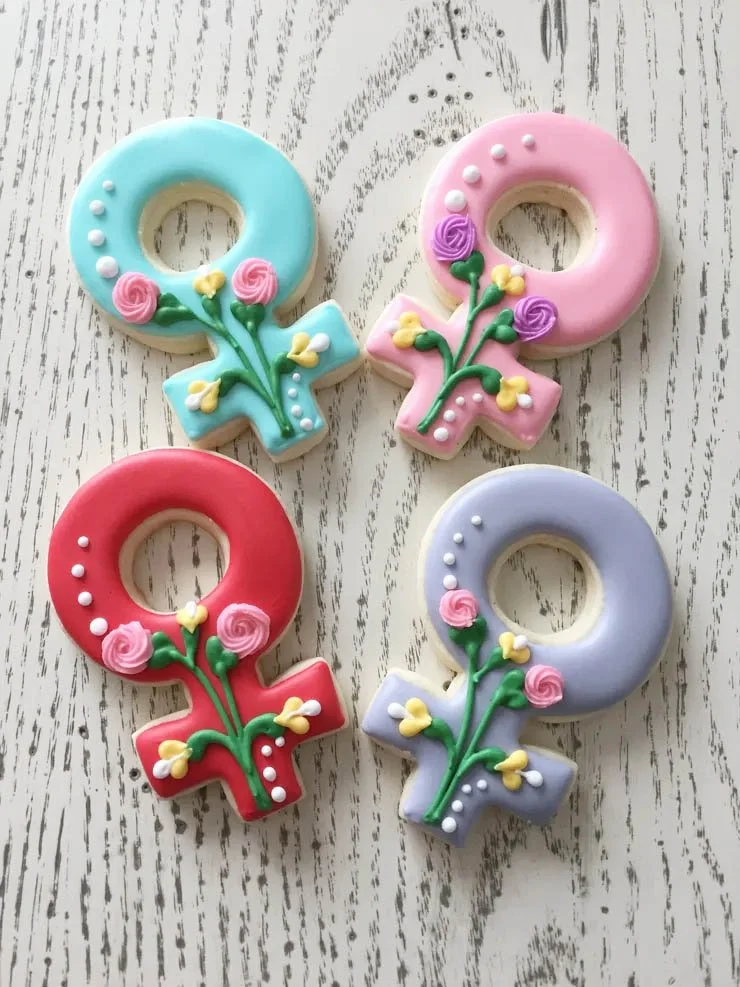 Decorated gender symbols cookies with floral designs, in pastel blue, pink, red, and lavender, placed on a white wooden surface.