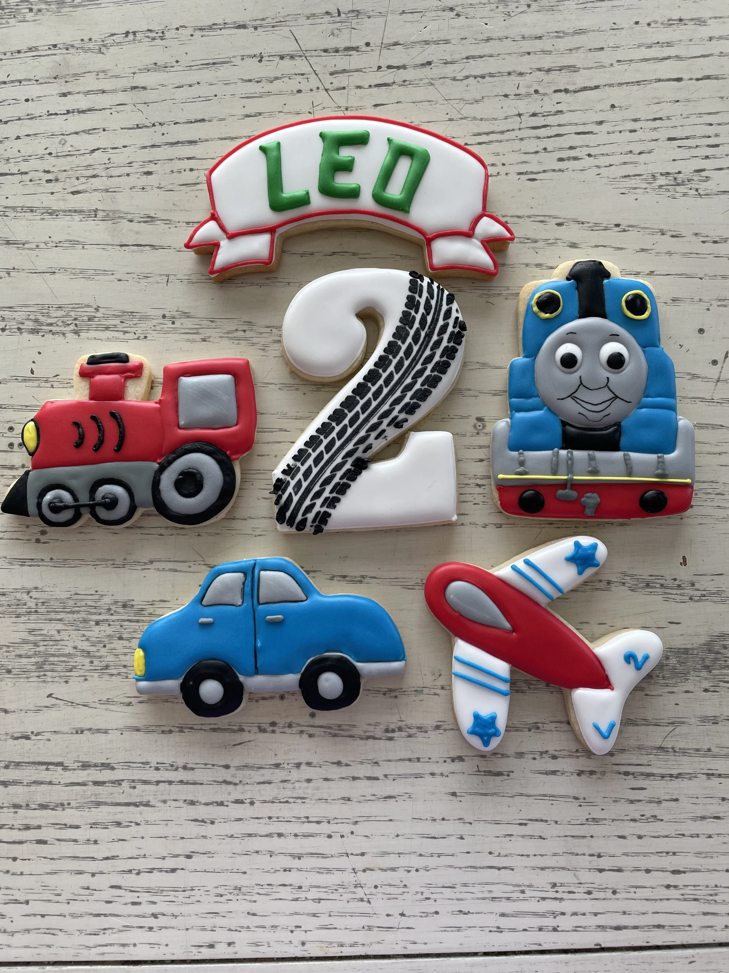 Decorated cookies themed around the children's TV show "Thomas & Friends," including a cookie with the name 'Leo,' a train engine cookie, a number 2 cookie, a car cookie, a plane cookie, and a smiling train face cookie.
