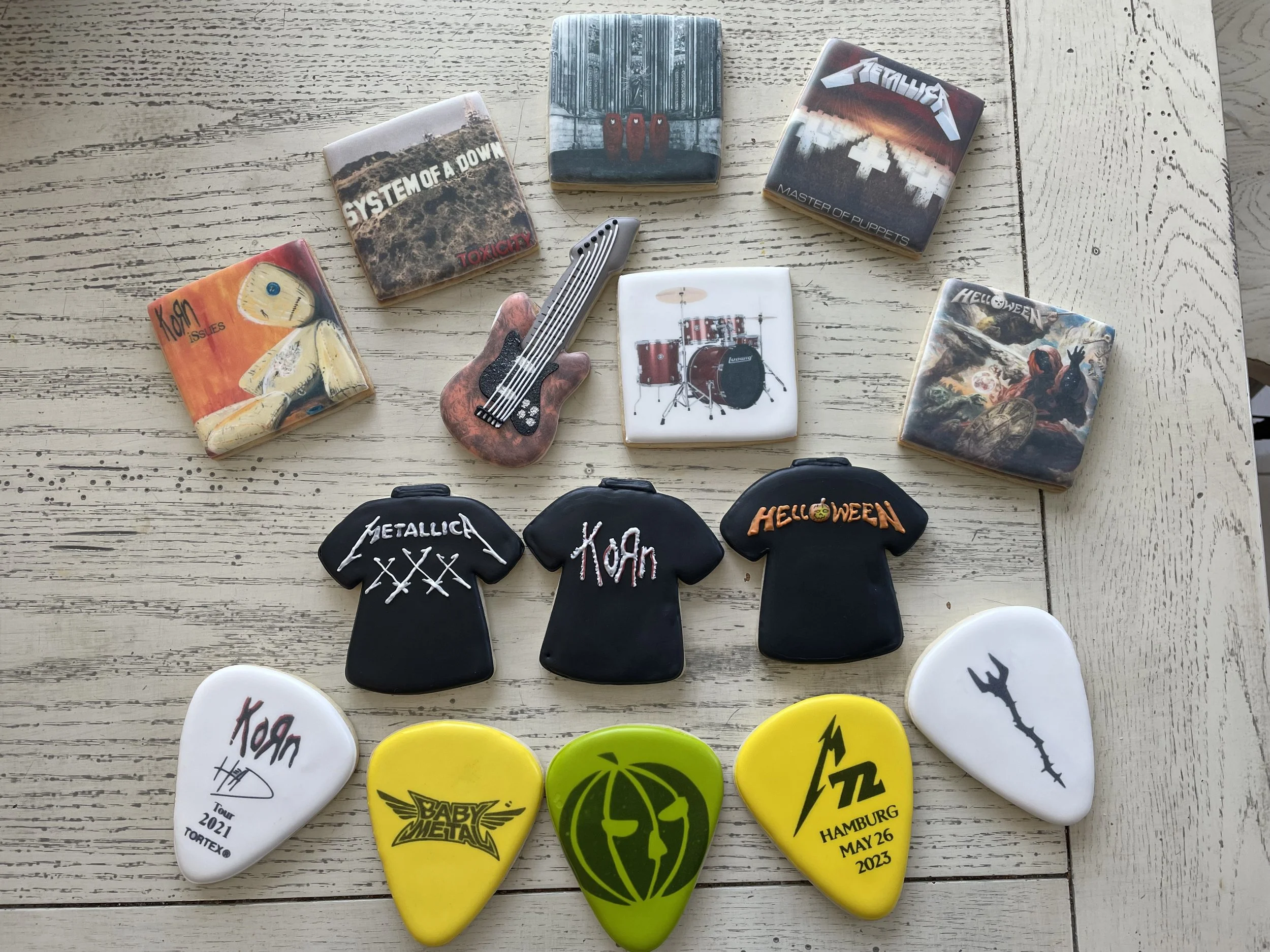 Collection of guitar picks, patches, and small band-themed items arranged on a wooden surface. The picks feature band logos like Metallica, Korn, Baby Metal, and Trivium, with some displaying tour dates and locations.