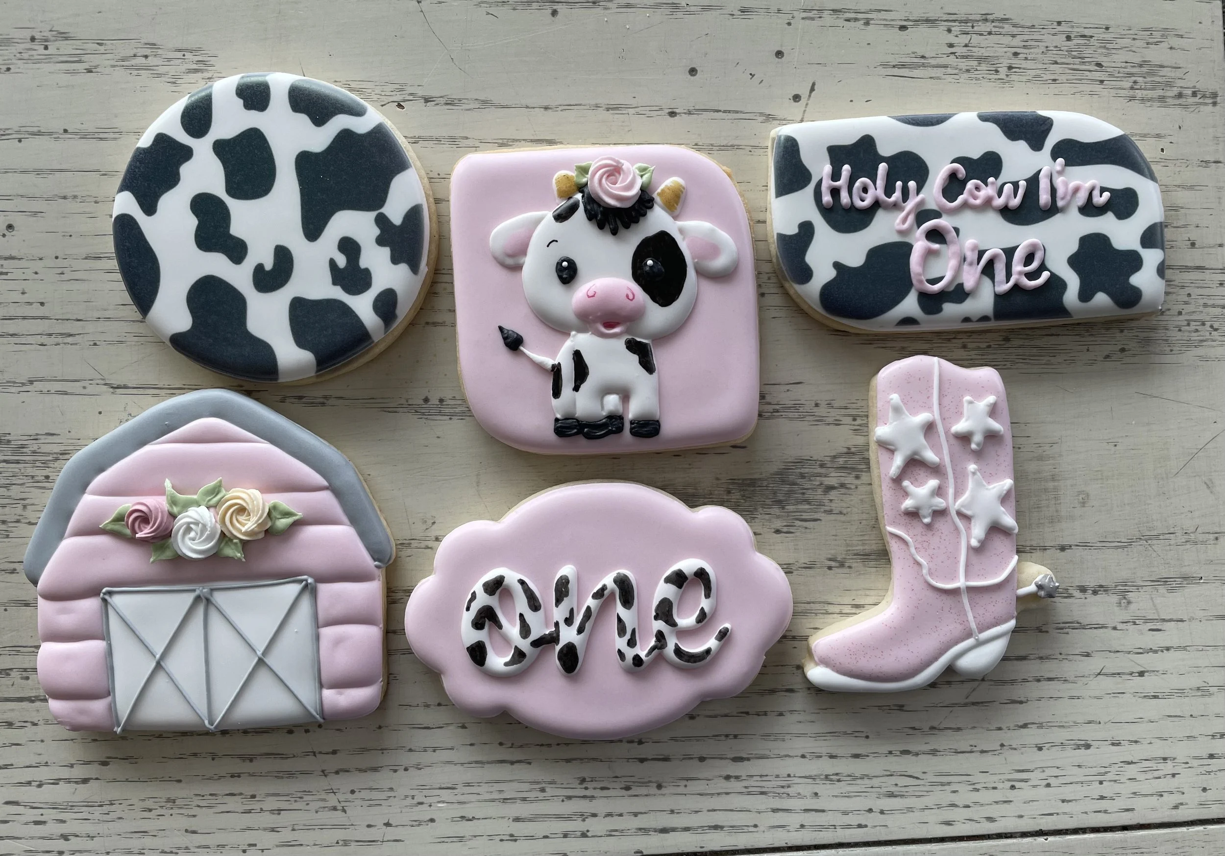 Decorative cookies with farm and love themes, including a black and white spotted cow, a pink pig, a cow print sign with the text "Holy Cow I'm One," a pink barn, the word "love" in cow print, and a pink cowboy boot with stars.