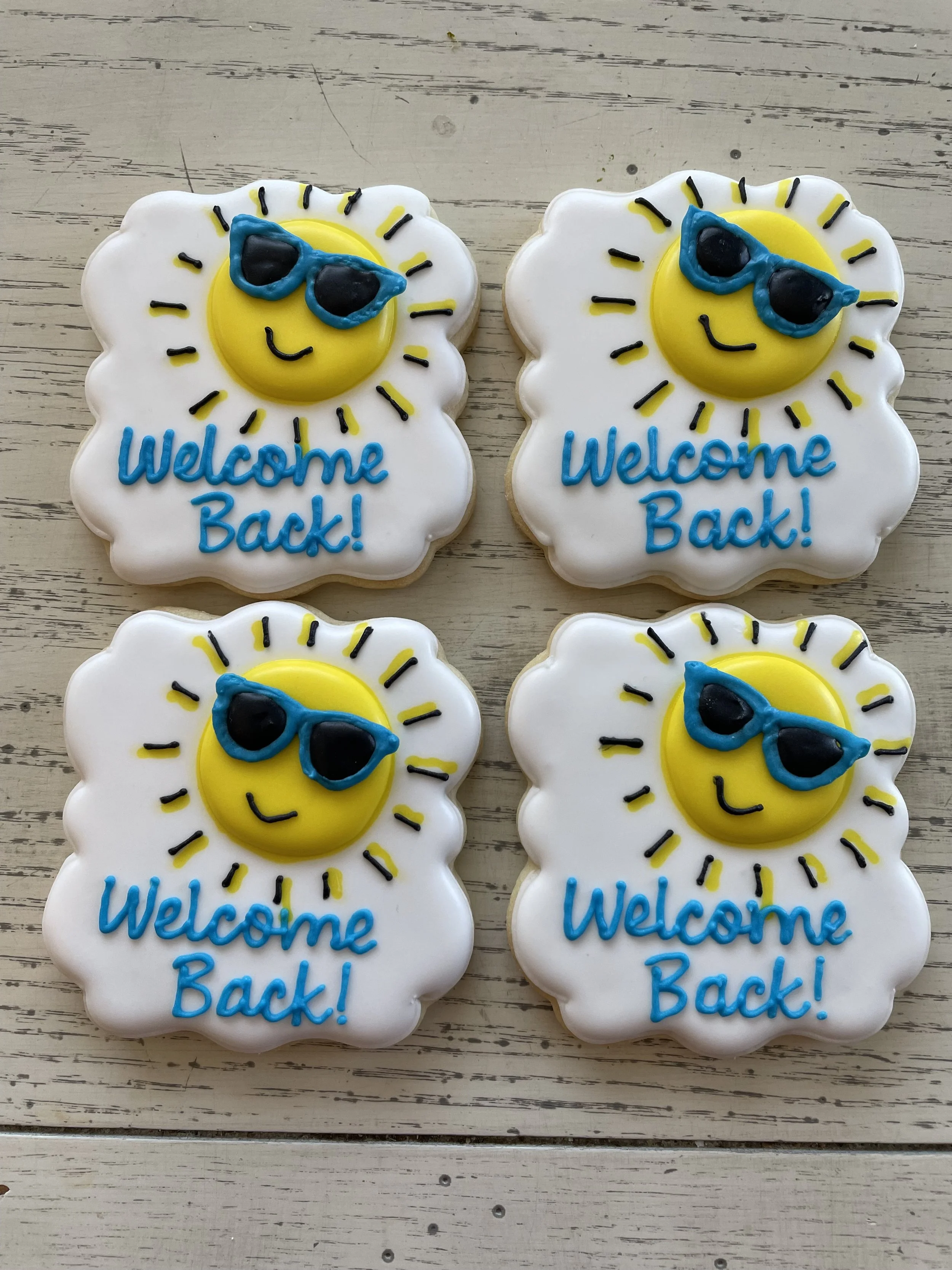Decorated cookies with a smiling sun wearing blue sunglasses, white icing background, and blue 'Welcome Back!' text.