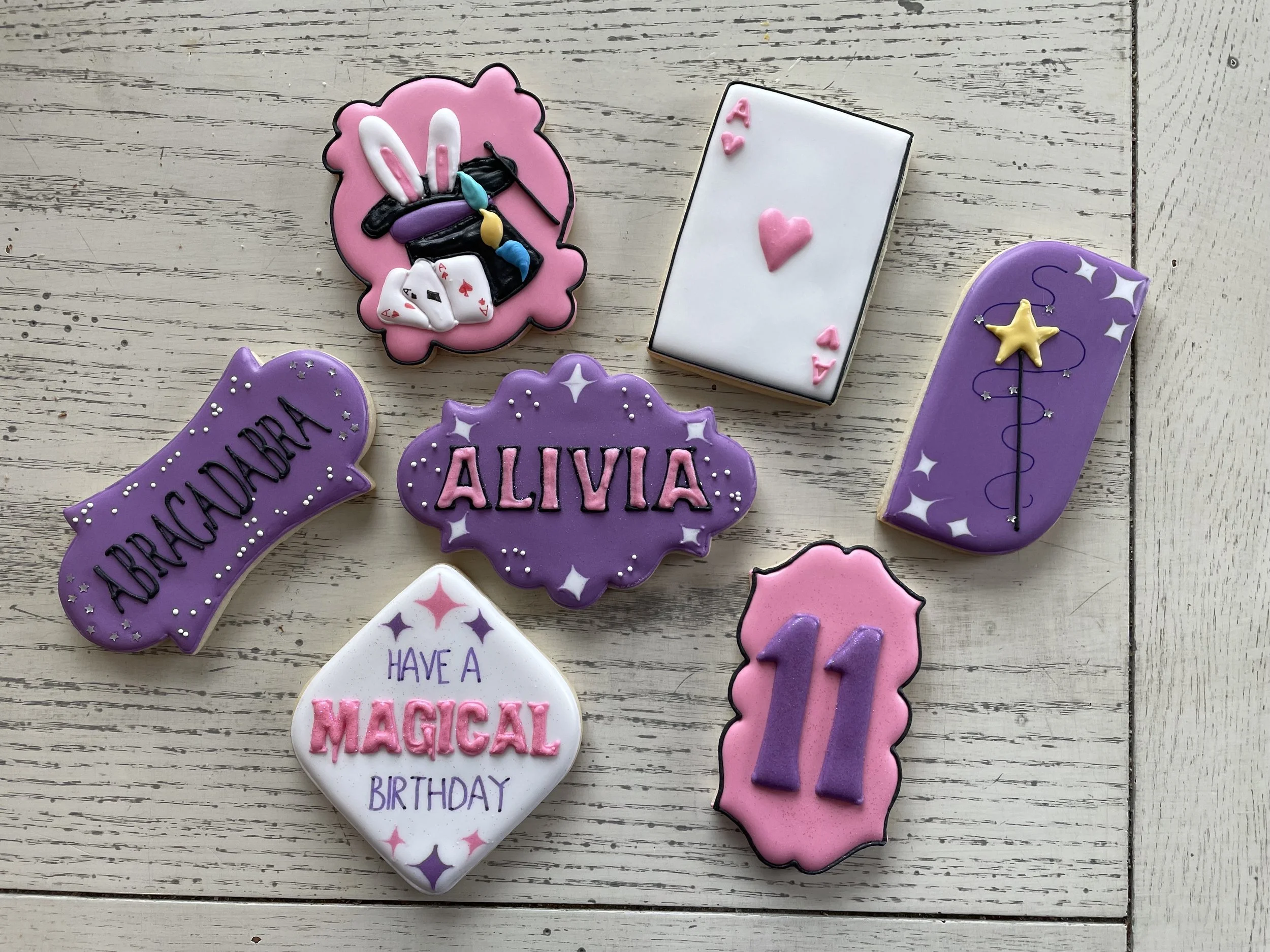 Decorated birthday cookies with magical and birthday theme, including a bunny with playing cards, a white rectangle with pink hearts, a purple cloud with "ALIVIA" written, a white diamond with "HAVE A MAGICAL BIRTHDAY," a purple moon with "ABRACADABR