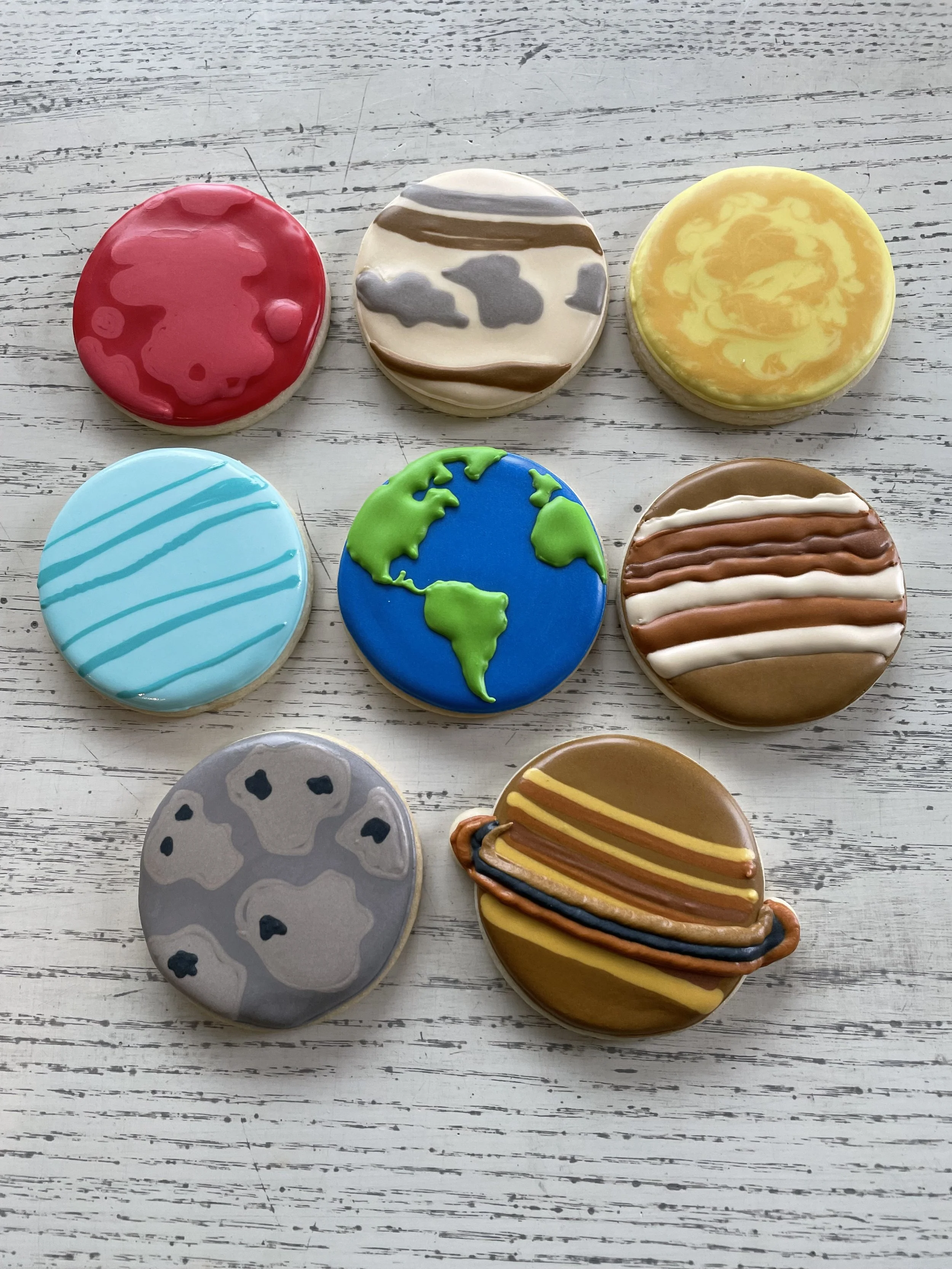 Set of nine astronomy-themed cookies decorated with colorful icing, featuring planets, Earth, stars, and space elements, arranged on a white wooden surface.